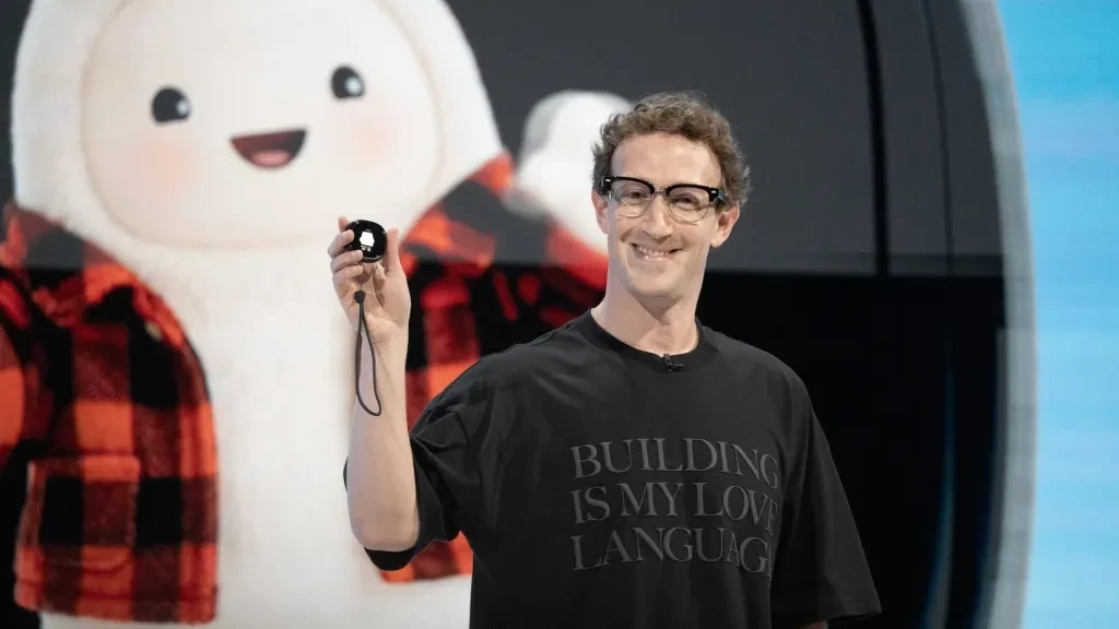 A presenter on stage holds up a small black device in front of a giant fluffy mascot on screen