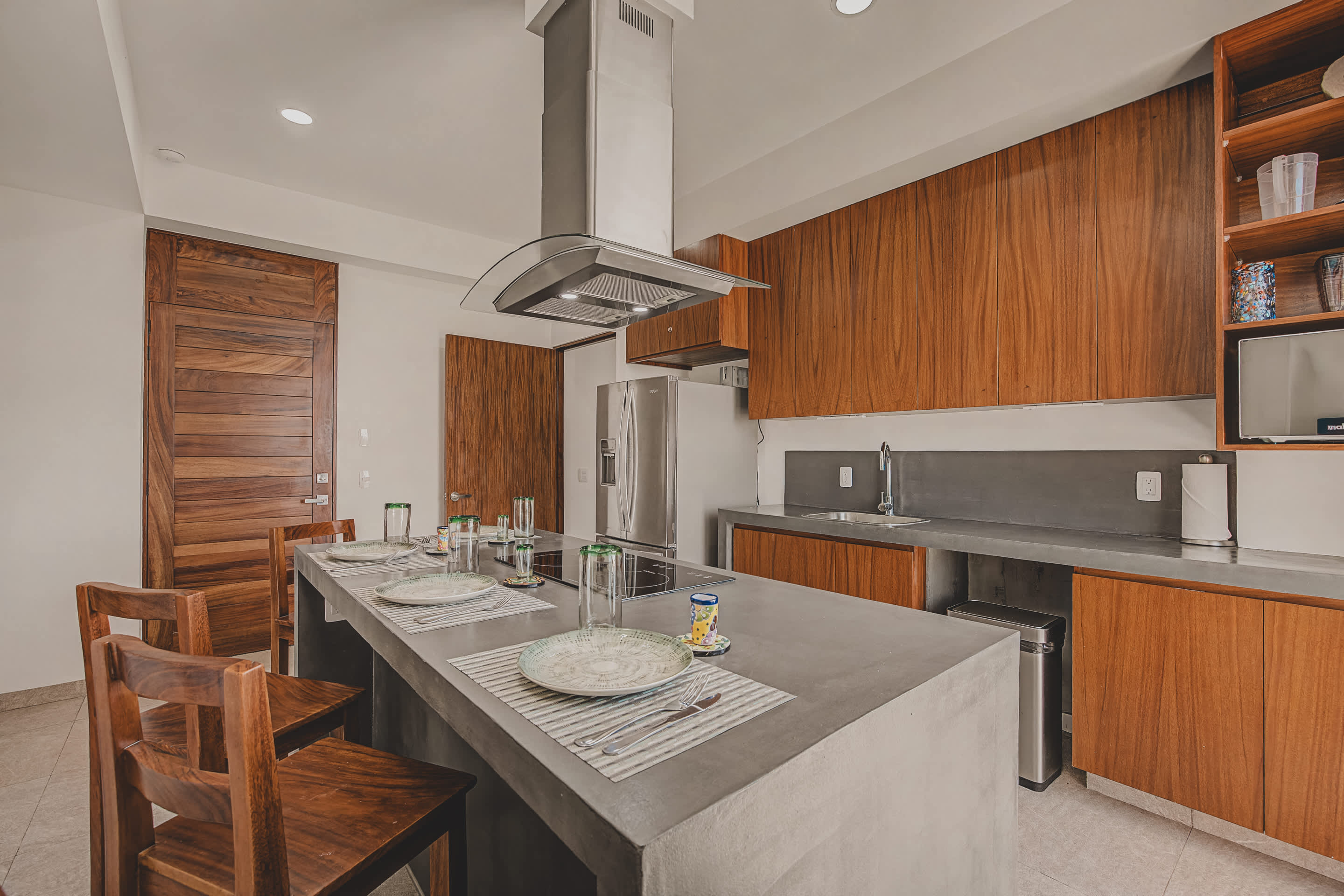 The spacious kitchen features a long dark grey island with an integrated cooktop and seating for multiple guests, surrounded by rich wooden cabinetry and modern stainless steel appliances, with a wooden door visible in the background.