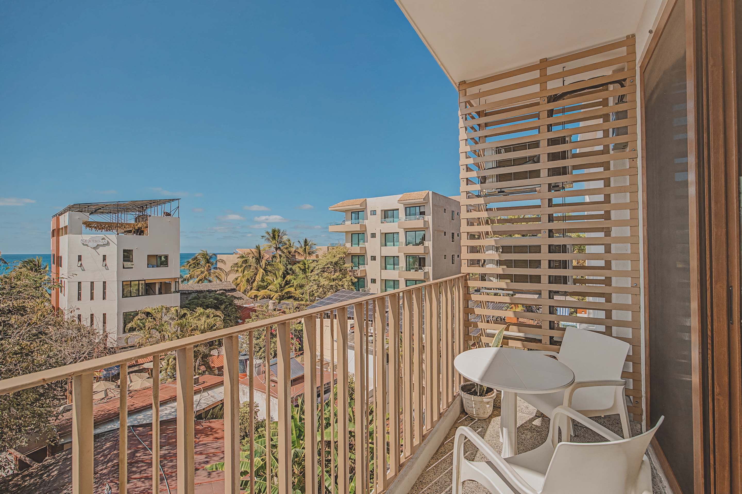 A bright balcony features a small white round table with two matching chairs, enclosed by a light-colored slatted railing, offering views of distant buildings and the ocean under a clear blue sky.