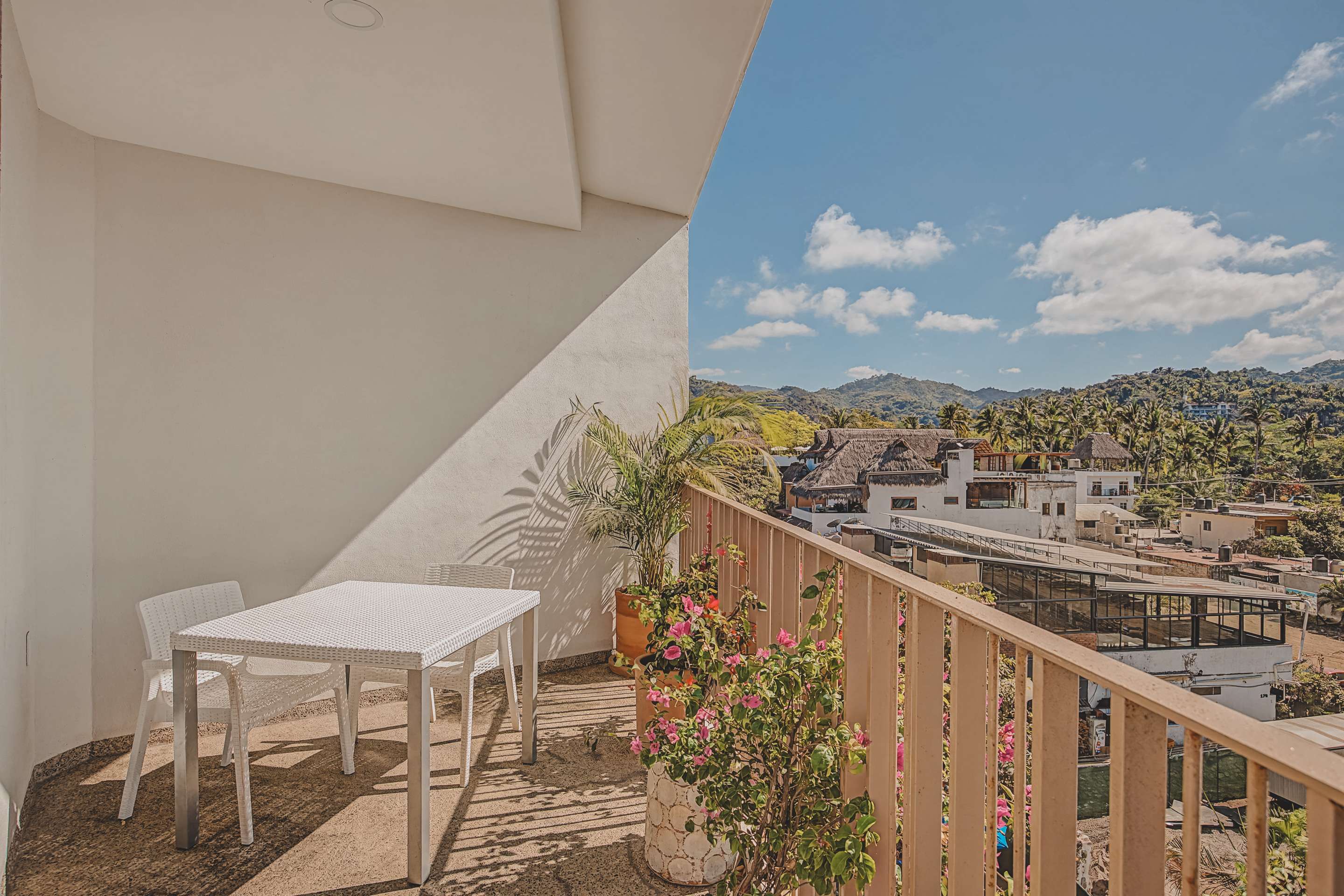 A spacious balcony features a white rectangular outdoor dining table with matching chairs, complemented by vibrant potted plants. The view extends to distant hills and a scattering of local rooftops.