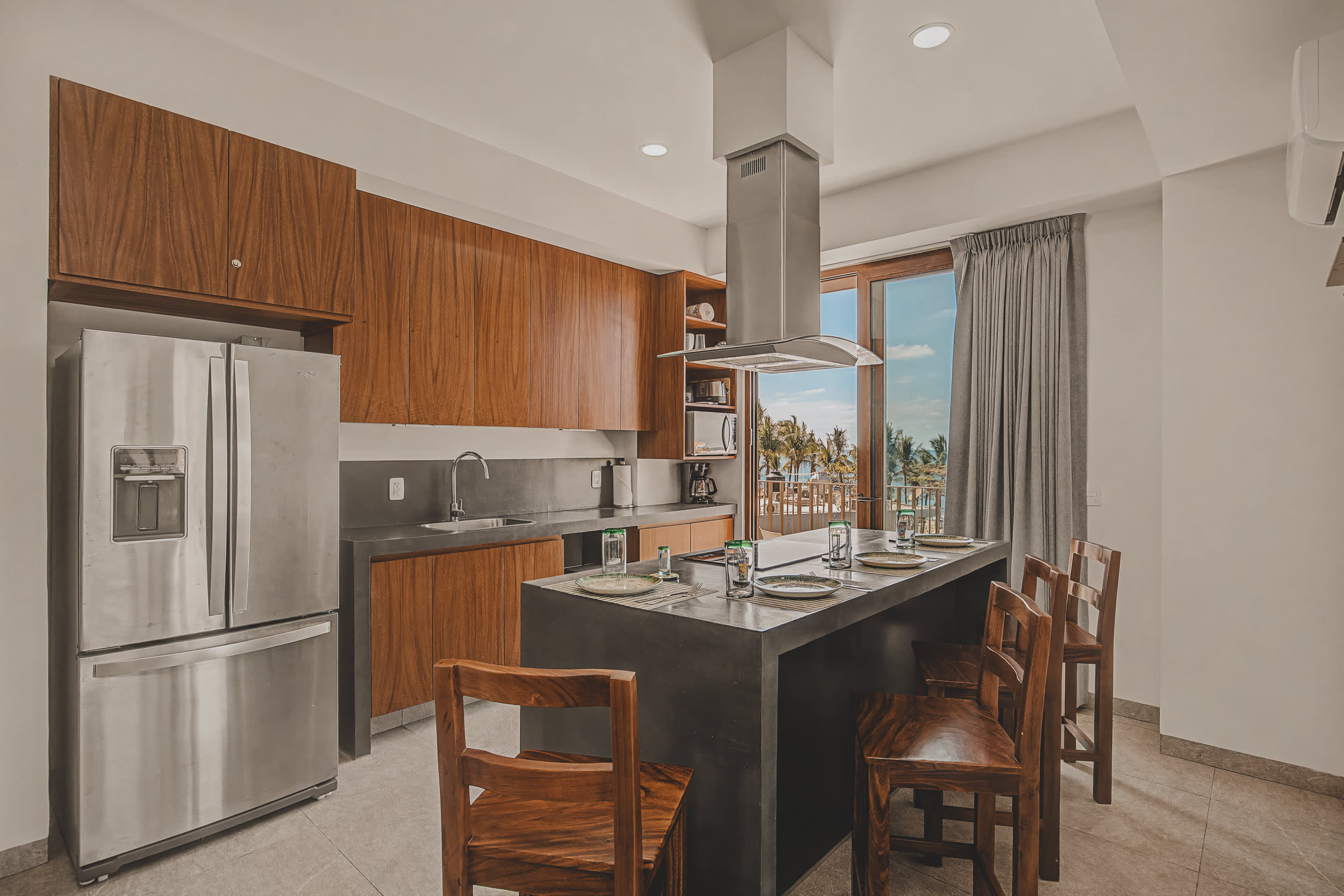 The contemporary kitchen showcases a large dark grey island with seating for four, surrounded by sleek wooden cabinets and stainless steel appliances, with a clear view of the ocean from the adjacent balcony.