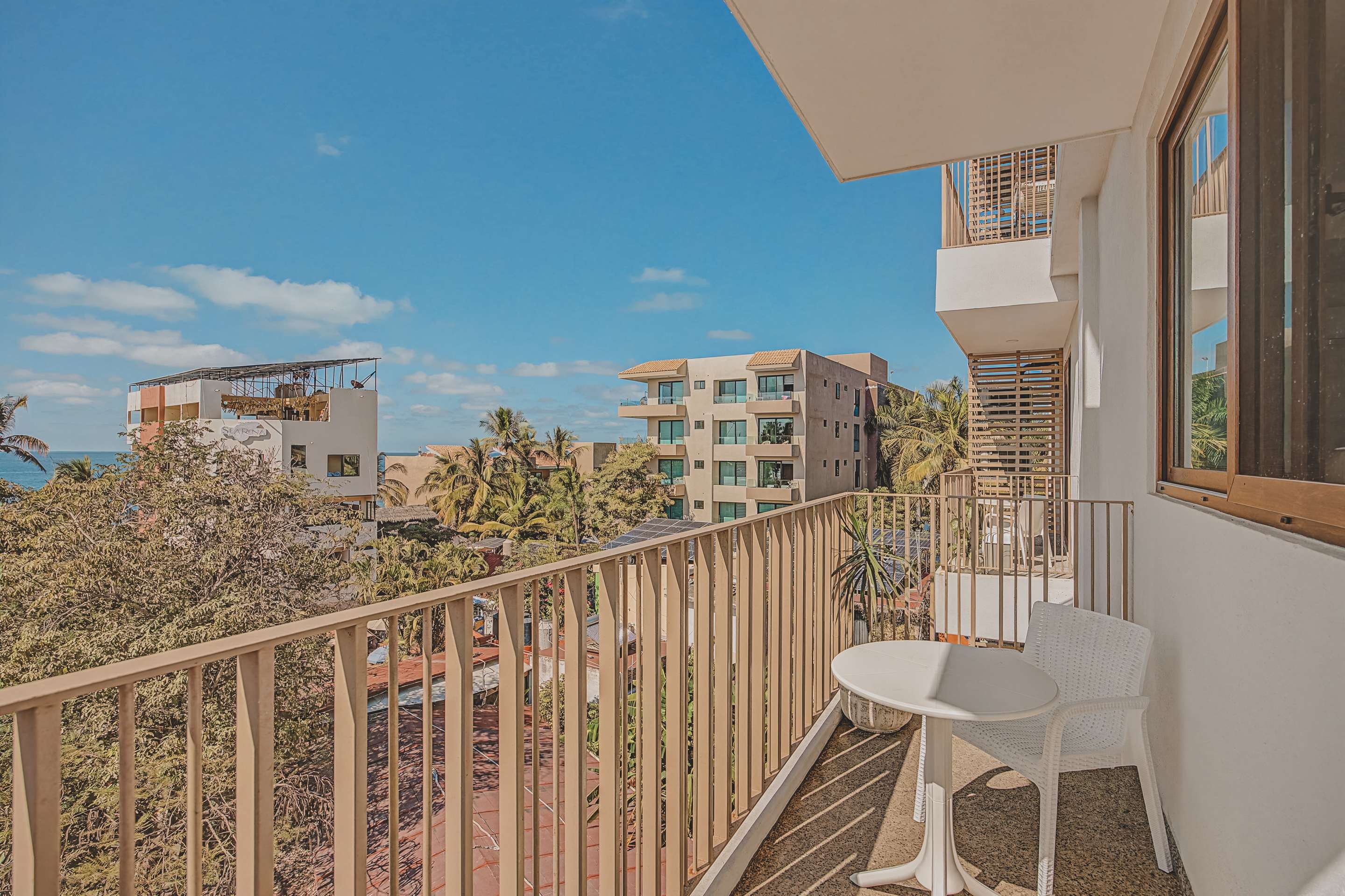 A bright balcony features a small white outdoor table and chair, offering a view of neighboring buildings, lush palm trees, and the distant ocean under a clear blue sky.