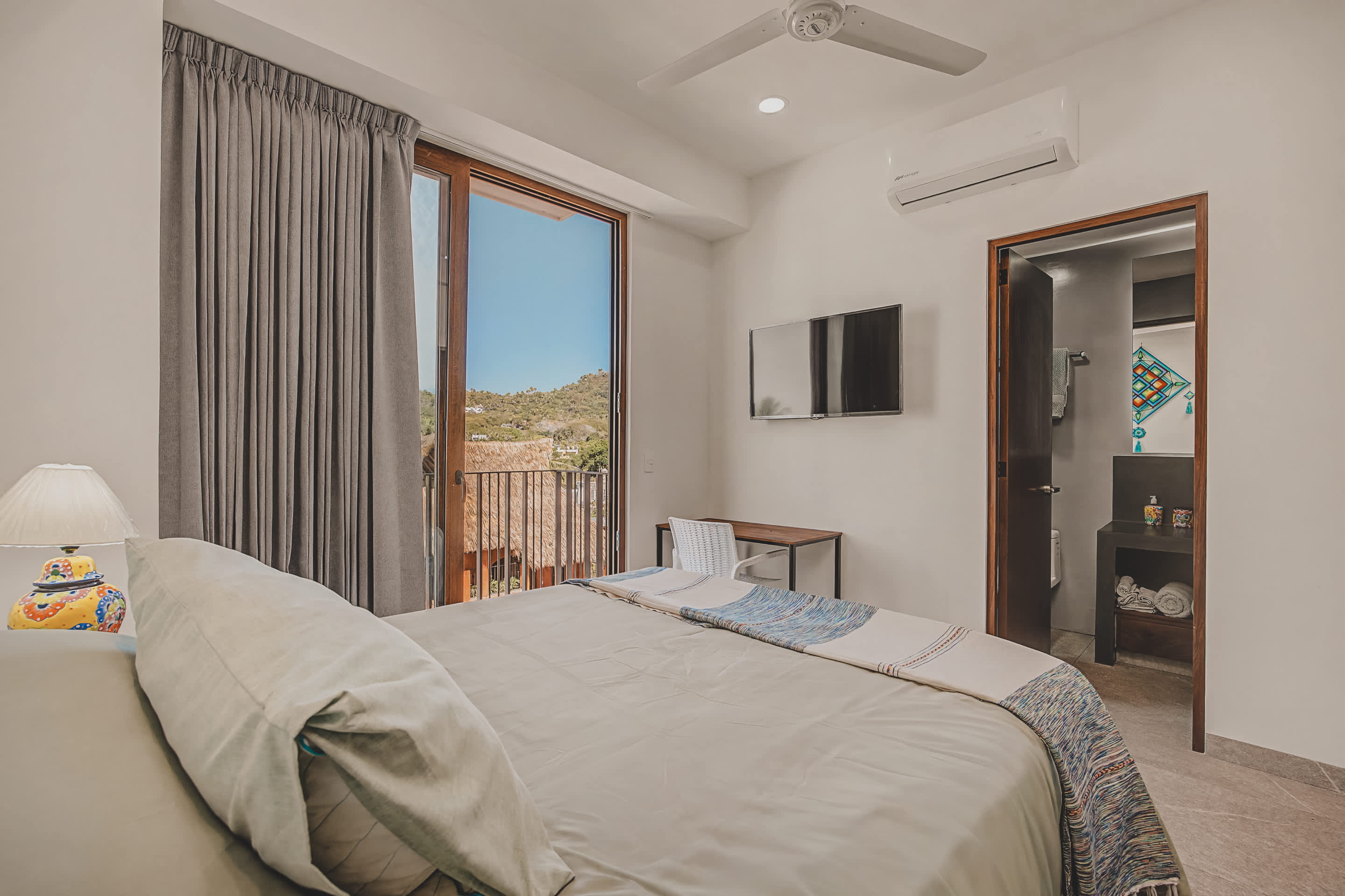 Bedroom with a queen bed, a wall-mounted TV, and an air conditioning unit above the doorway. A glass door leads to a balcony with views of distant hills, and an open doorway reveals a modern ensuite bathroom.