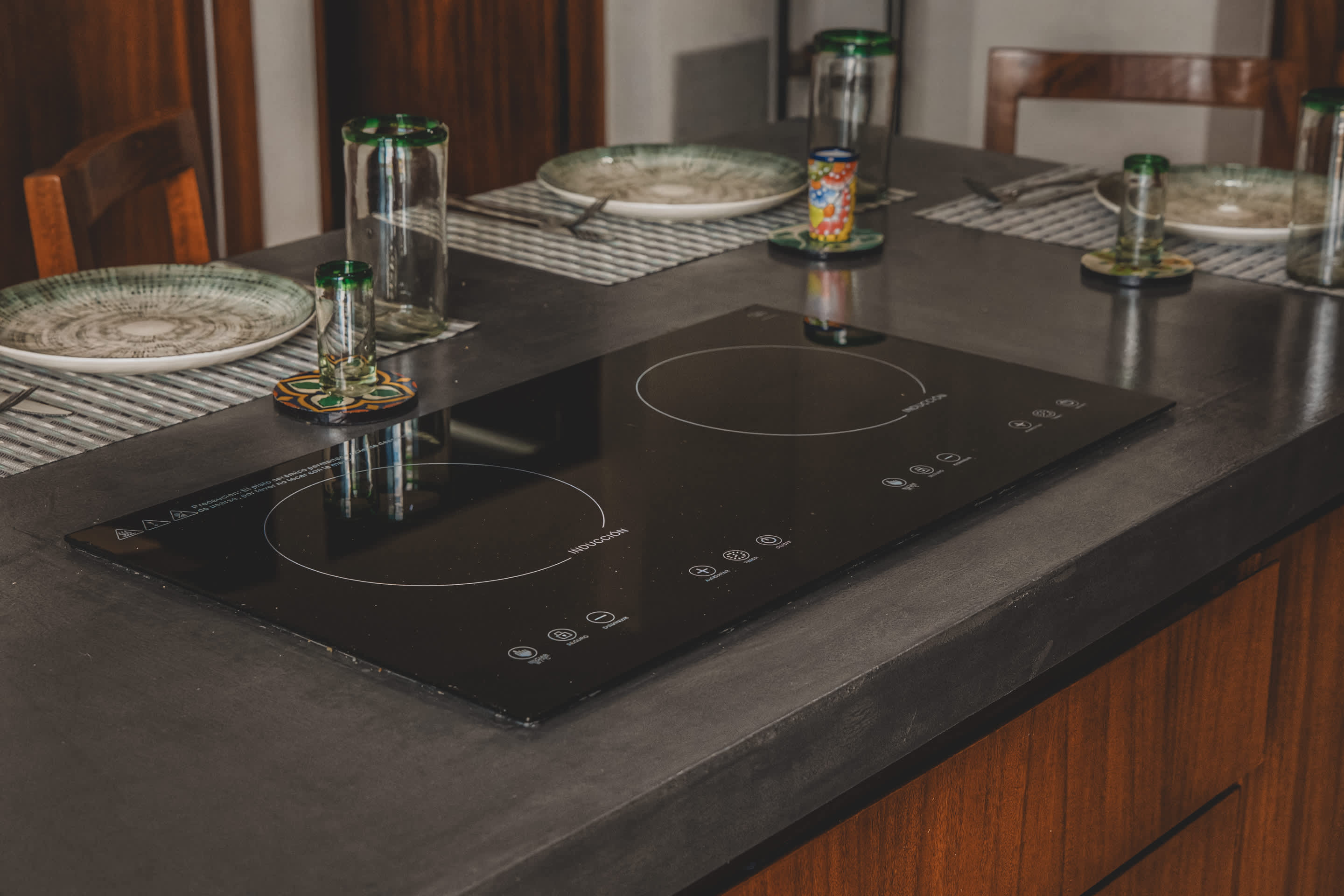 Sleek black induction cooktop integrated into a dark concrete countertop, surrounded by dining place settings with green and white plates and tall glasses.