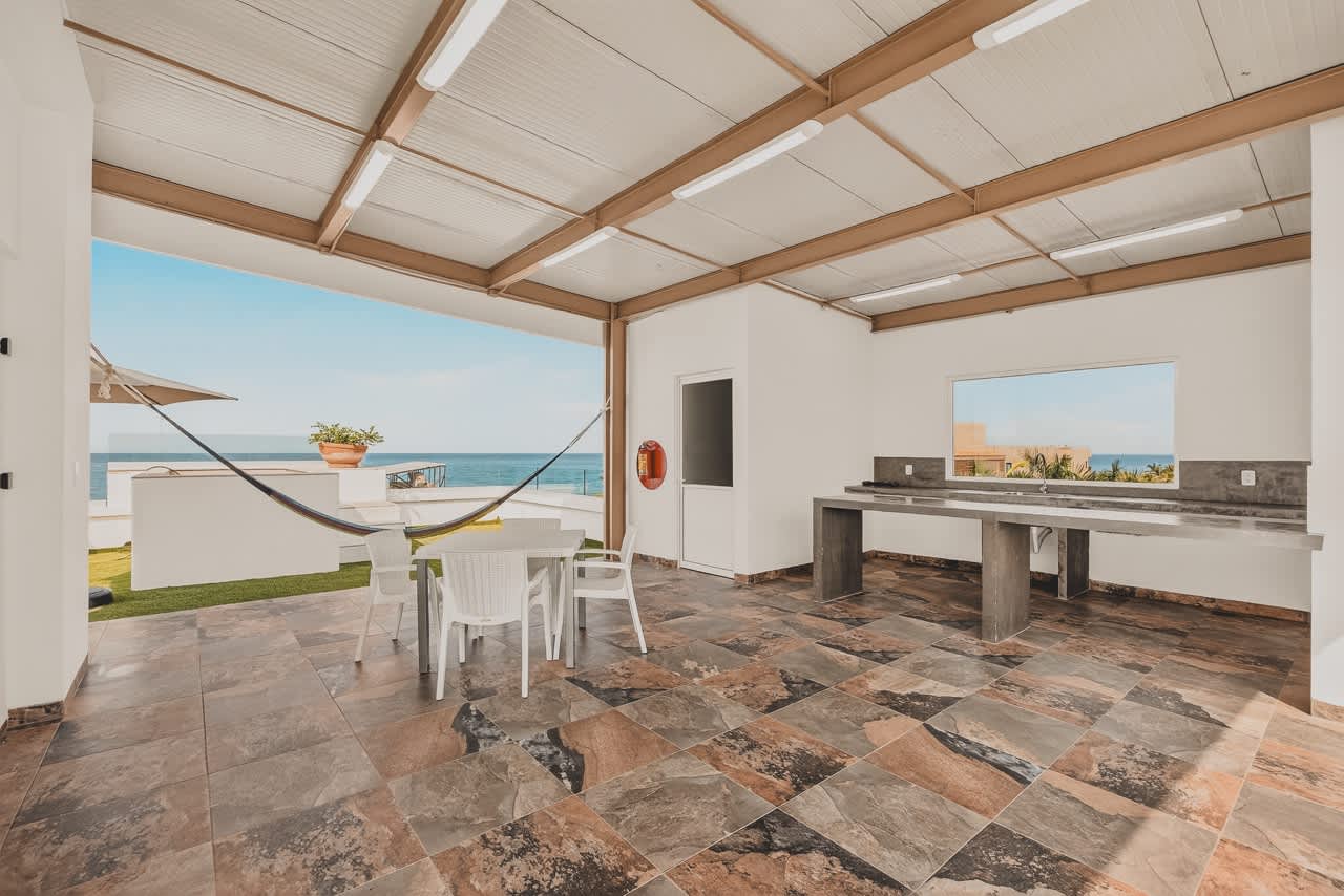 Spacious covered outdoor area combining a kitchen with concrete counters and a dining space featuring a white plastic table and chairs. The multi-toned tiled floor extends to an open view of the ocean and a grassy area with a hammock.