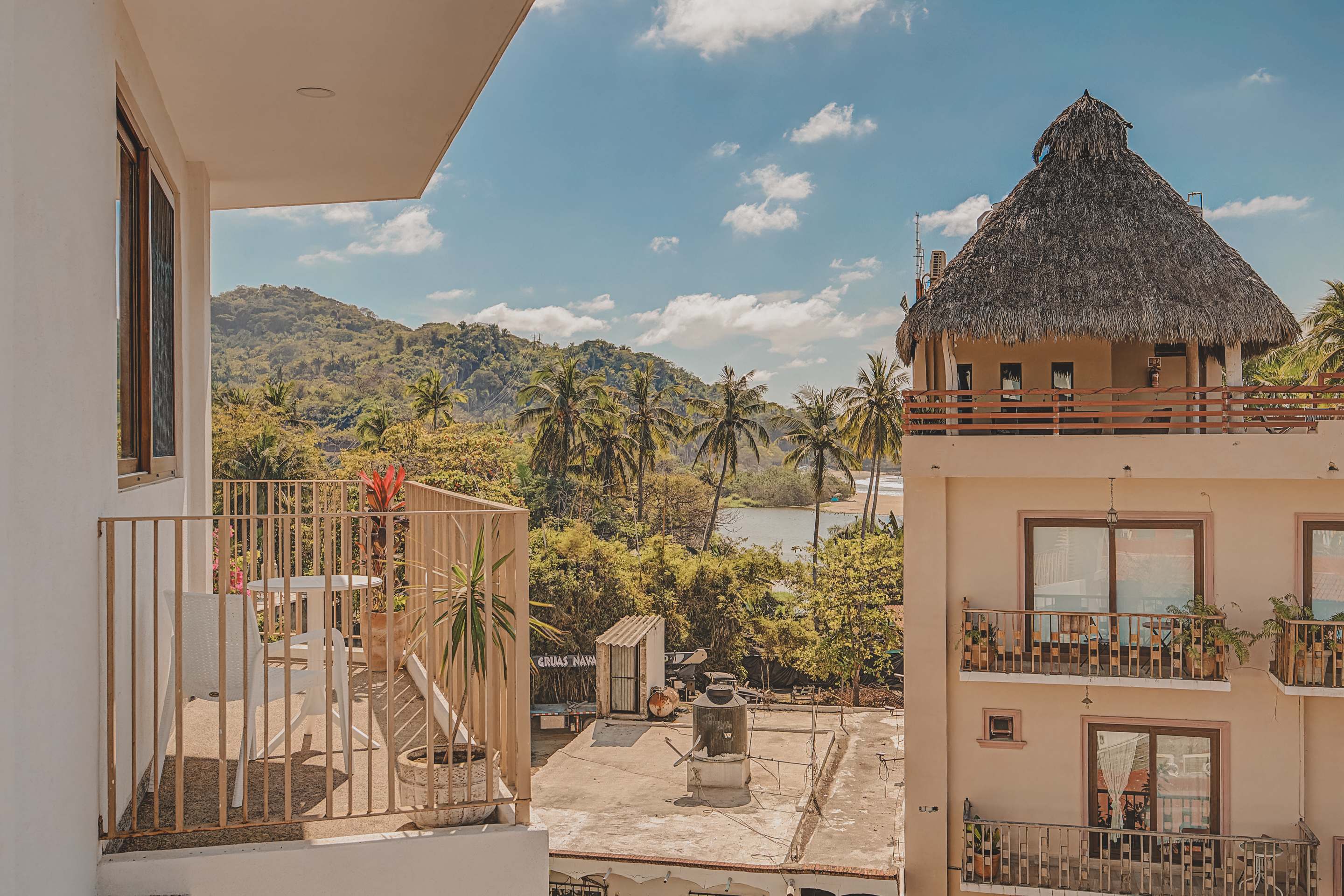 A picturesque view reveals a tranquil river or lagoon winding through dense green foliage, backed by rolling hills and mountains, with a distinctive building featuring a thatched roof in the foreground.