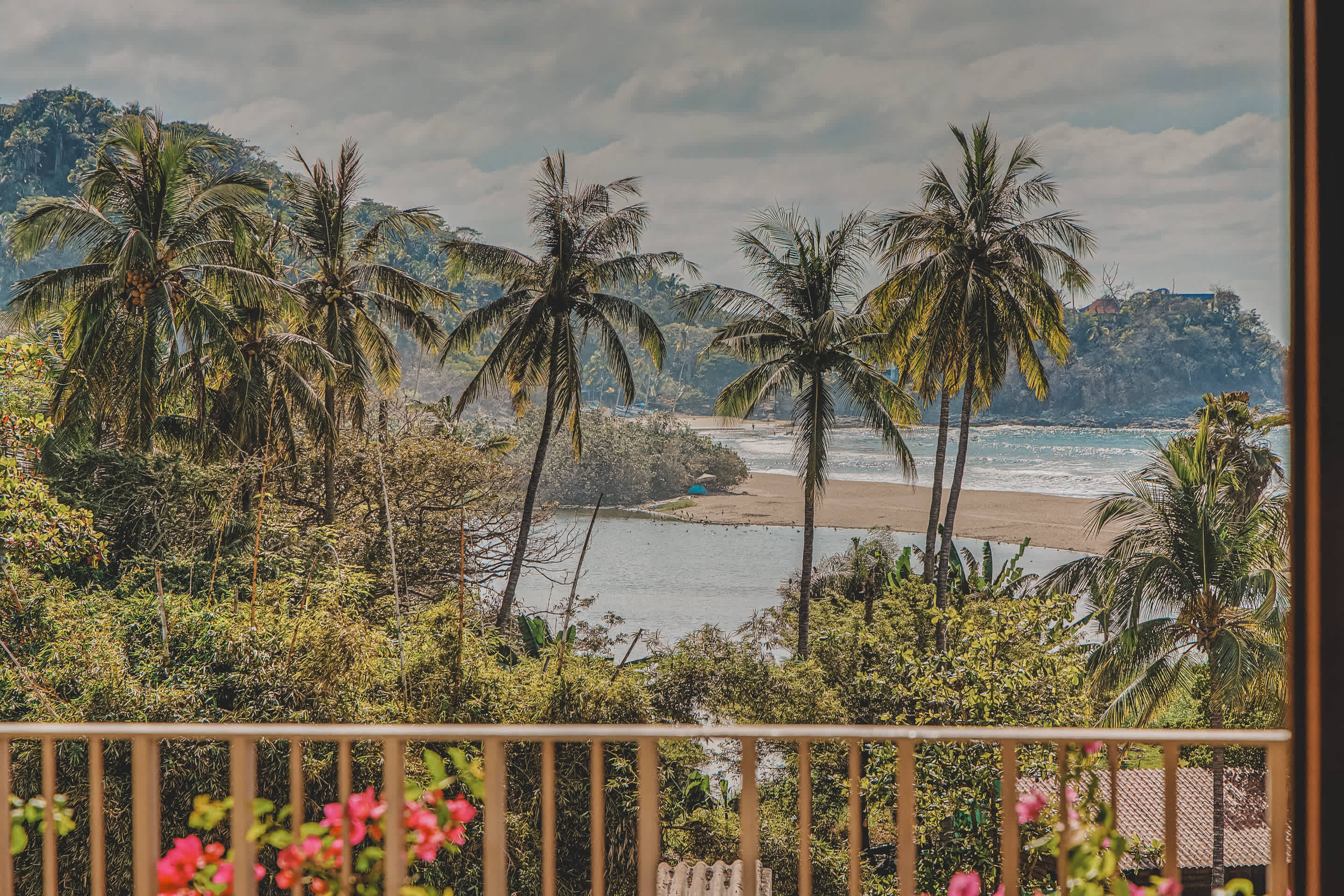 Picturesque view from a balcony overlooking a tranquil bay with a sandy beach, surrounded by dense tropical foliage and numerous palm trees under a cloudy sky.