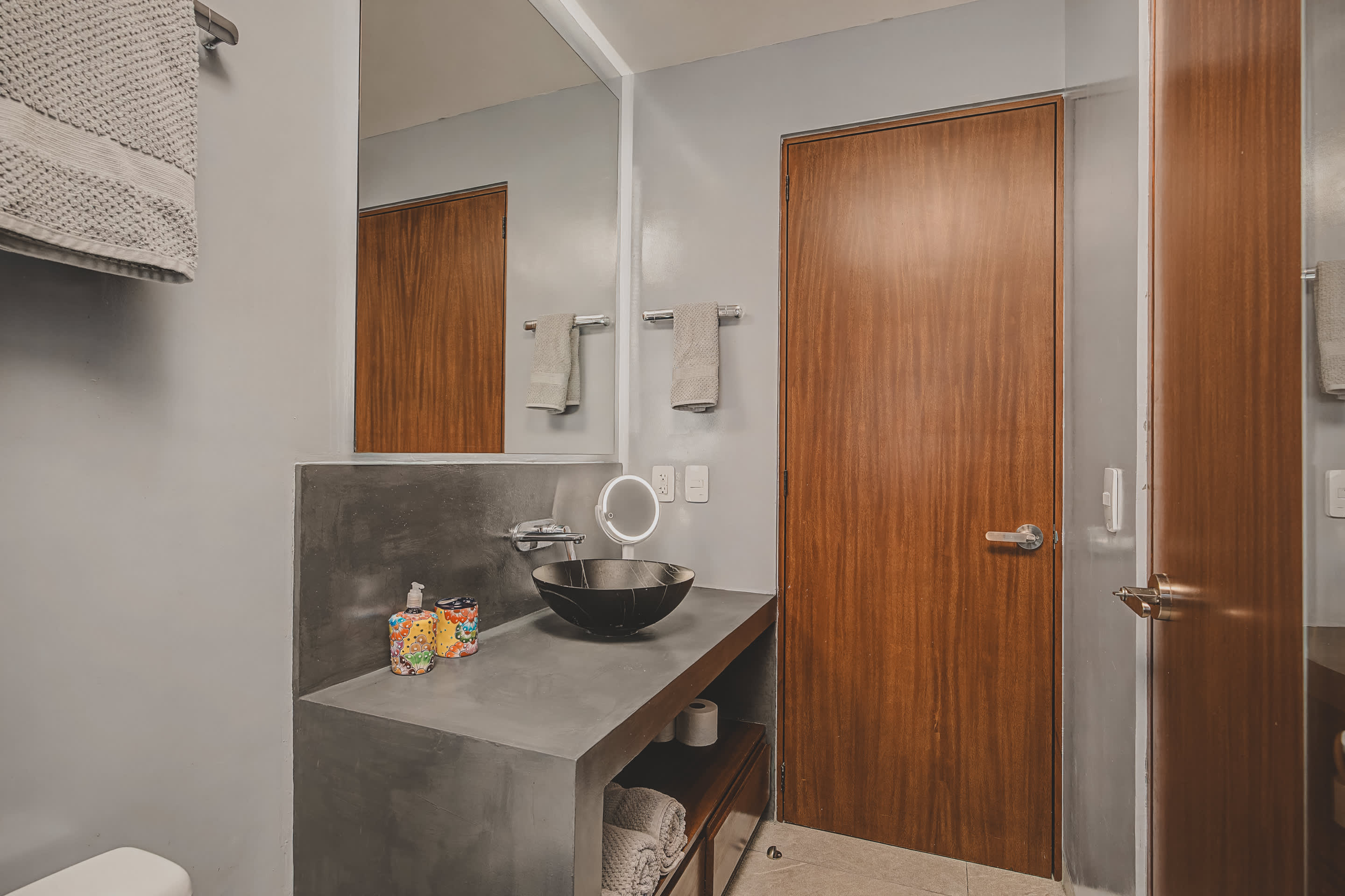 Close-up view of the modern bathroom vanity, featuring a black vessel sink set into a dark concrete countertop, beneath a large mirror. Rolled towels are neatly stored on a shelf below the sink, and a wooden door is visible to the right.