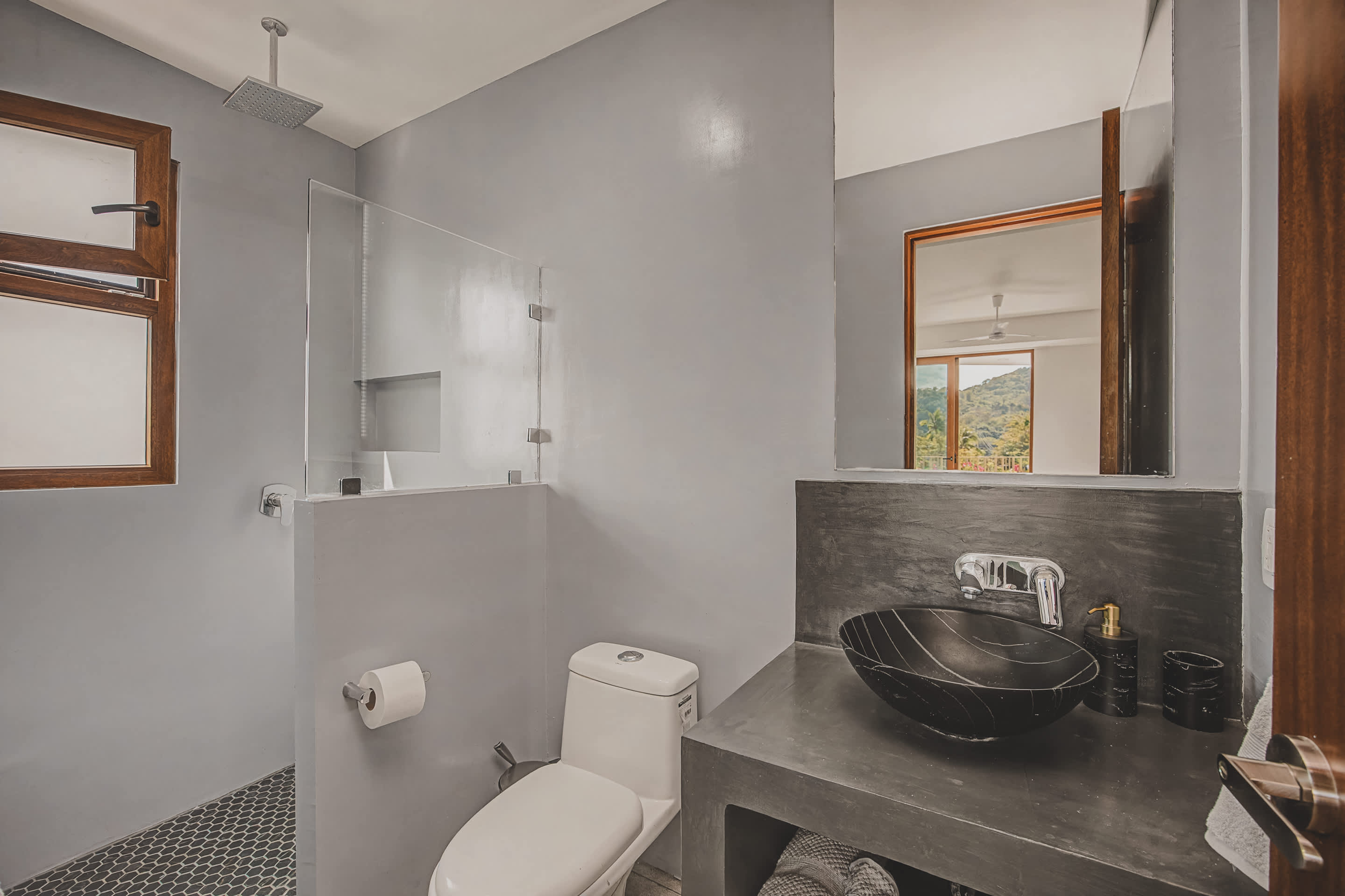 Modern bathroom featuring light gray walls, a walk-in shower with a rain showerhead and hexagonal tile floor, and a dark concrete vanity with a black vessel sink. A wooden-framed window provides natural light.
