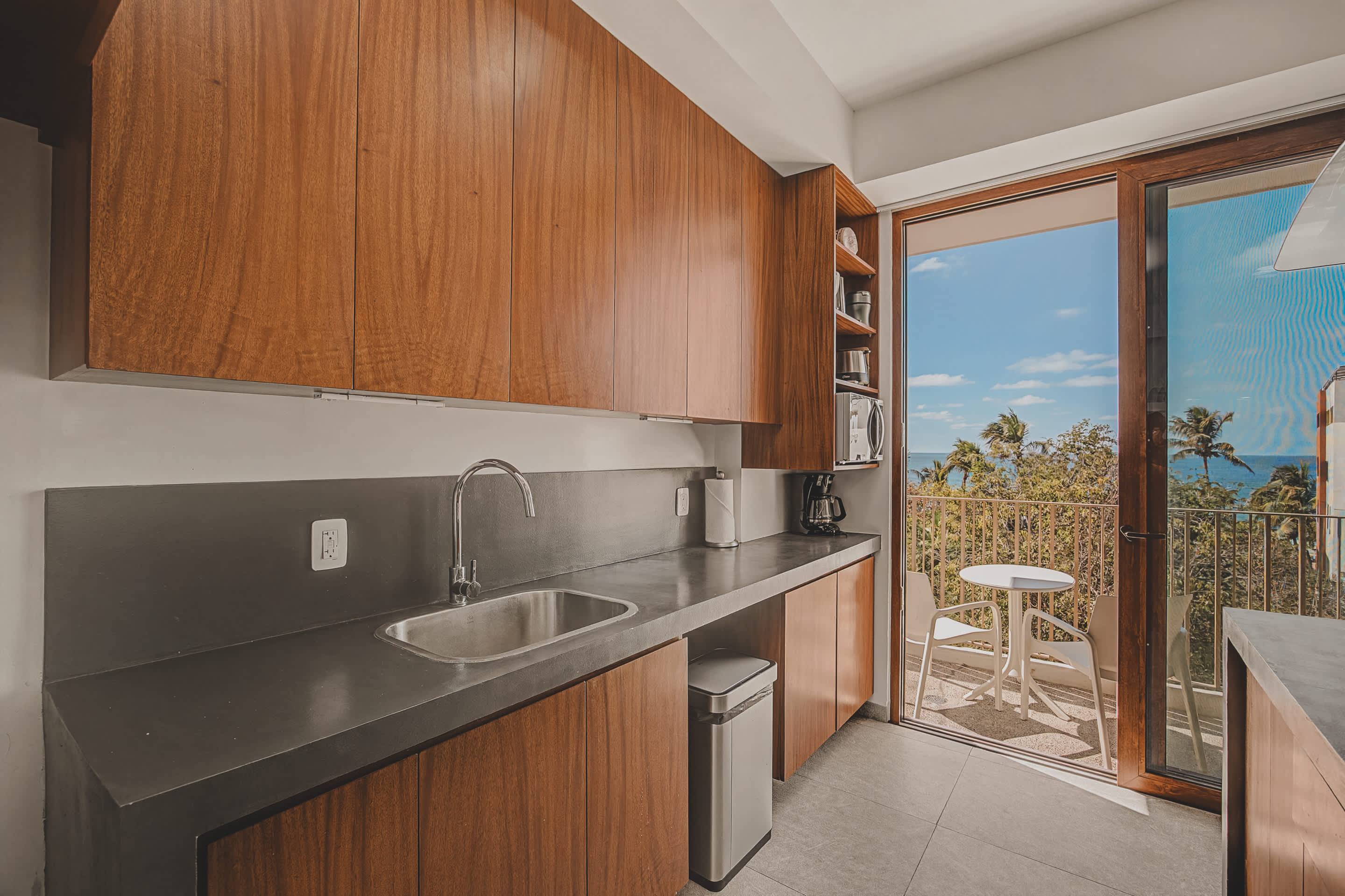 A detailed view of the kitchen counter highlights a stainless steel sink set into dark grey countertops, with ample wooden cabinetry above and below, and a sliding glass door leading to a bright balcony.