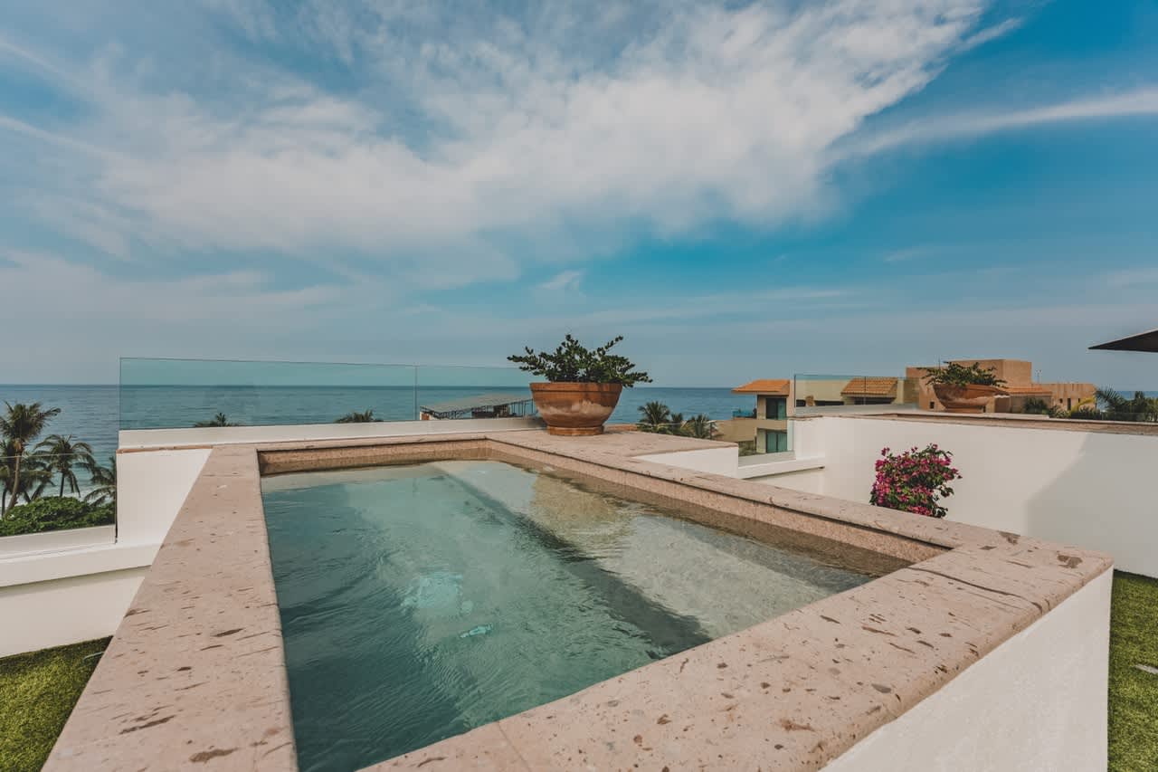 A serene rooftop plunge pool with stone edges, overlooking the vast blue ocean and distant buildings. The clear water reflects the bright sky, creating a tranquil and inviting atmosphere.