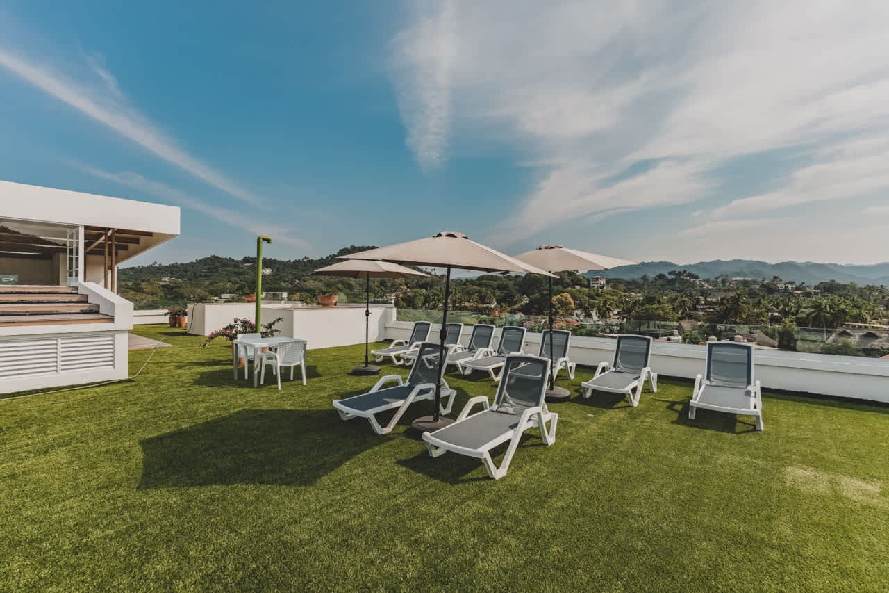 Expansive rooftop terrace covered in artificial green turf, furnished with numerous white lounge chairs and two large beige umbrellas for shade. A small dining set is also present, offering views of distant green hills under a bright blue sky.