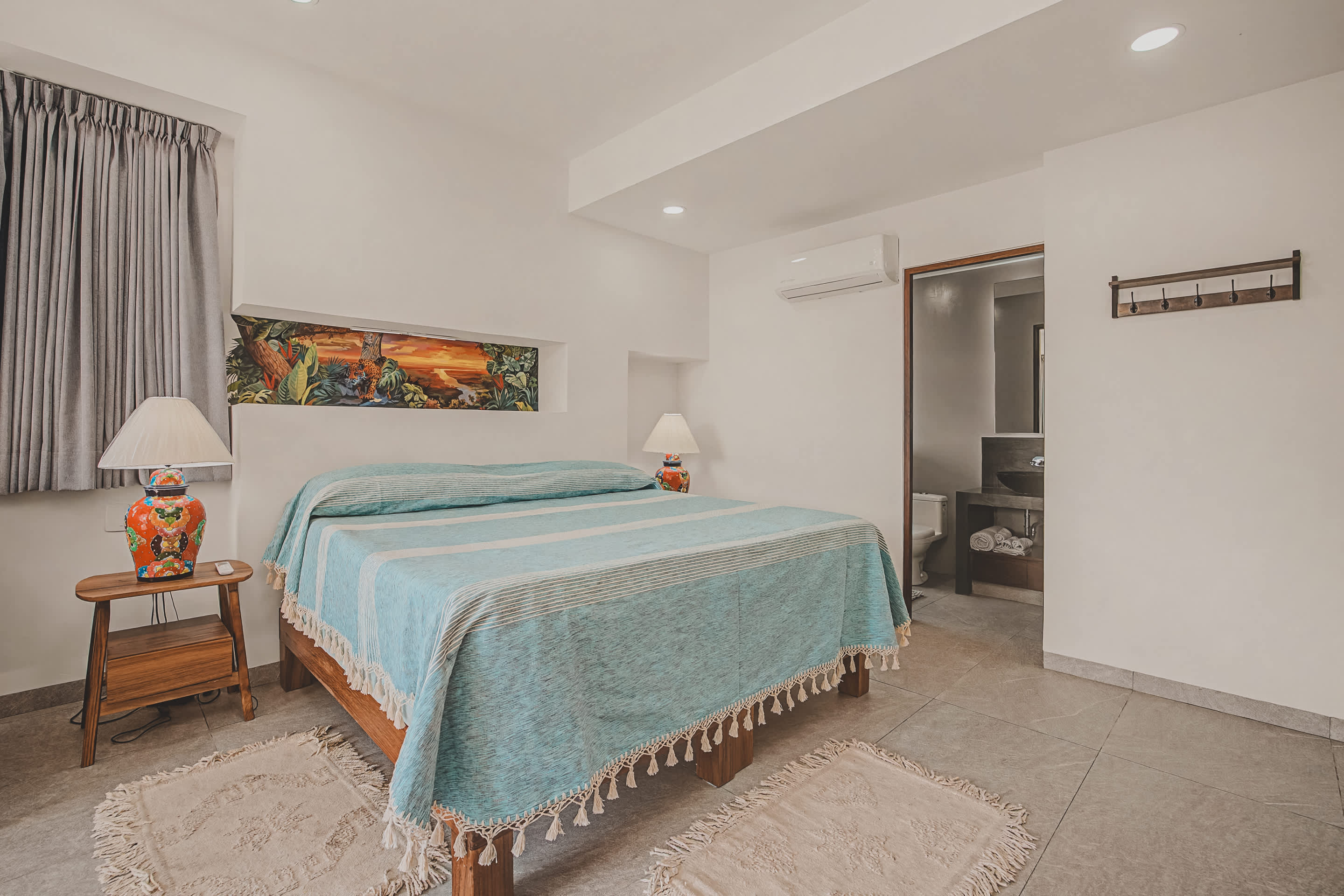 The bedroom's sleeping area features a bed with a light blue striped cover and fringe, flanked by wooden bedside tables with vibrant, hand-painted lamps. An open doorway leads to an adjacent bathroom.