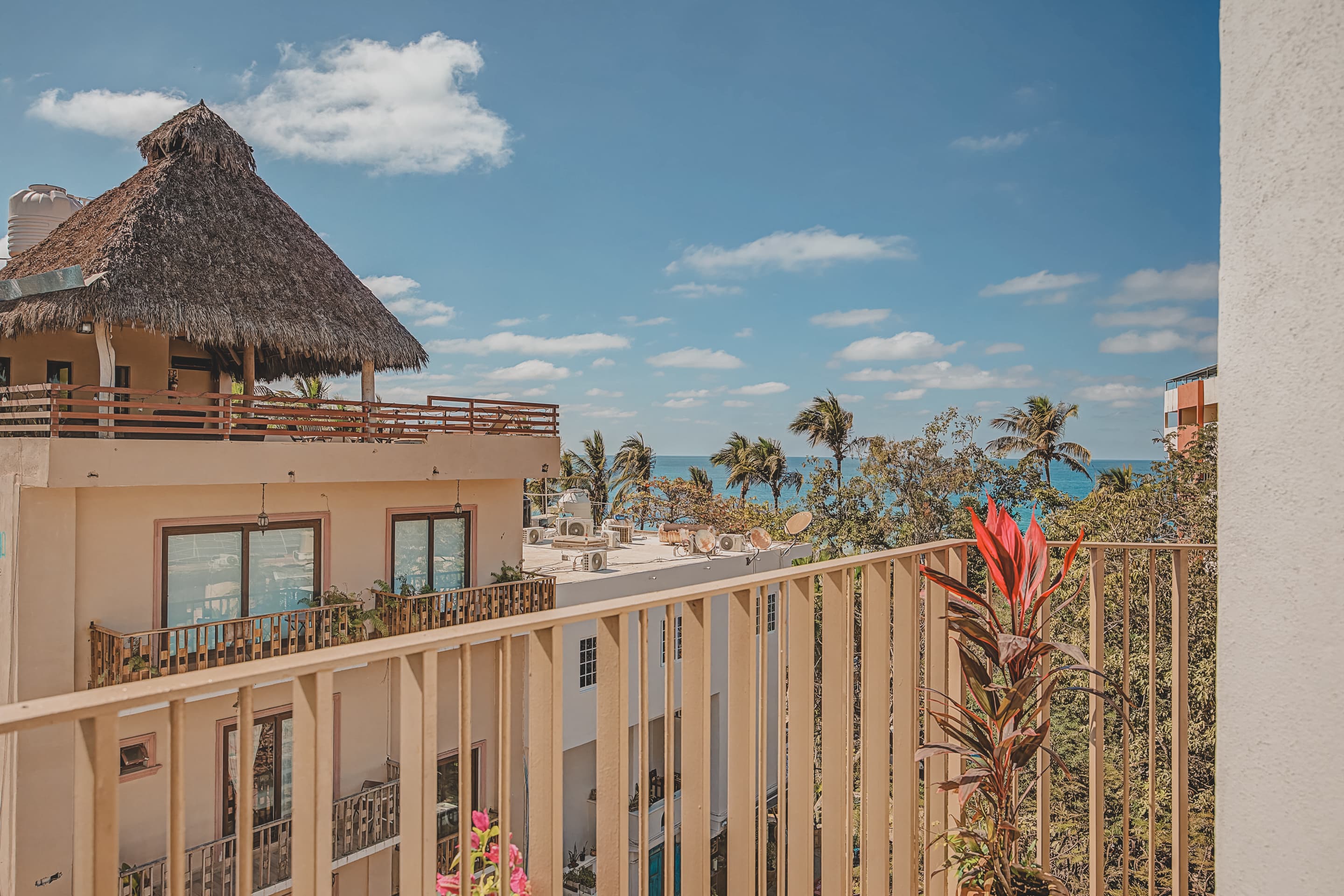 A balcony view captures a nearby building with a distinctive thatched roof and wooden railings, with glimpses of the blue ocean and palm trees in the background under a bright sky.
