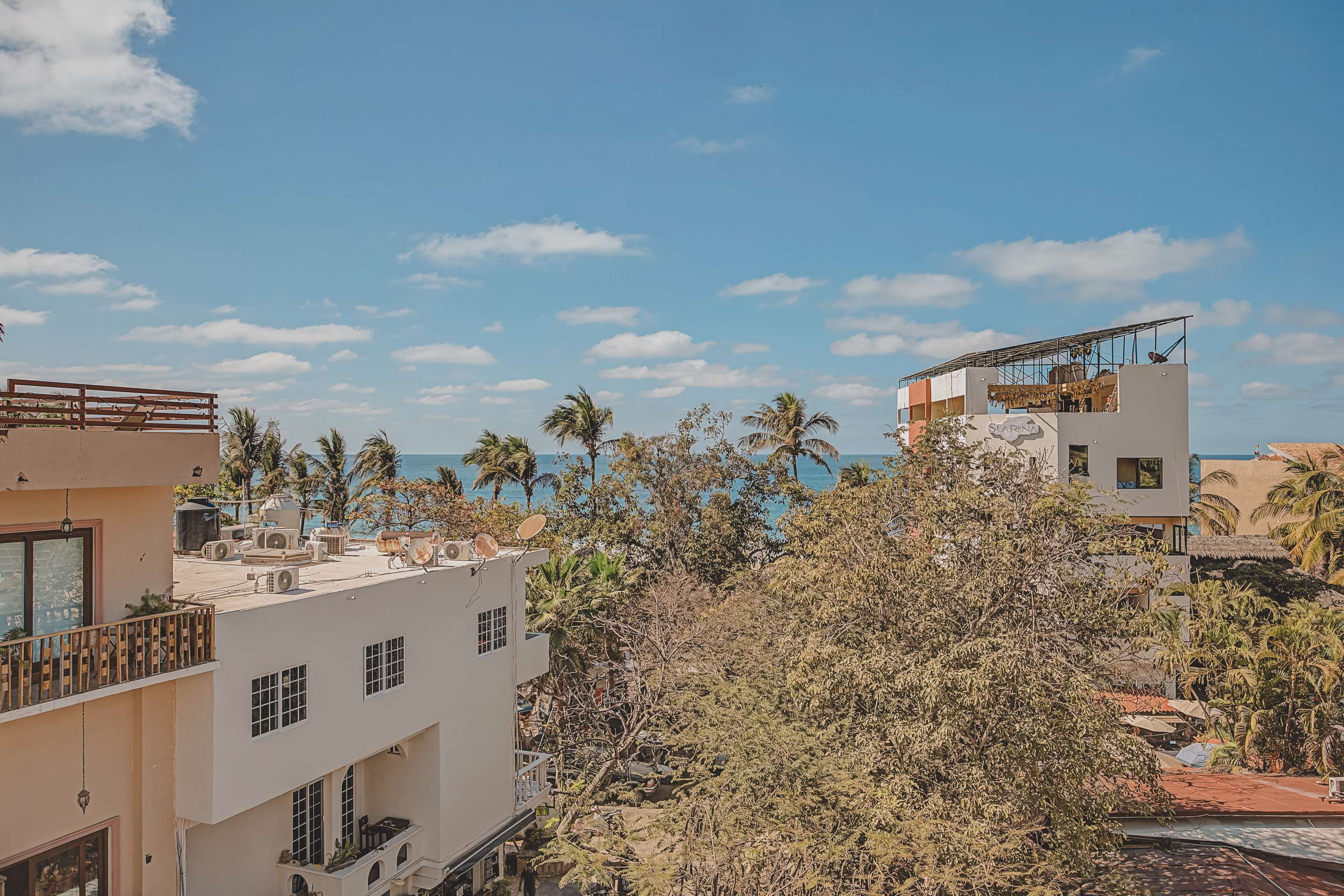 This balcony view overlooks several white buildings with flat roofs and air conditioning units, with the sparkling blue ocean and a line of palm trees visible in the distance under a clear sky.