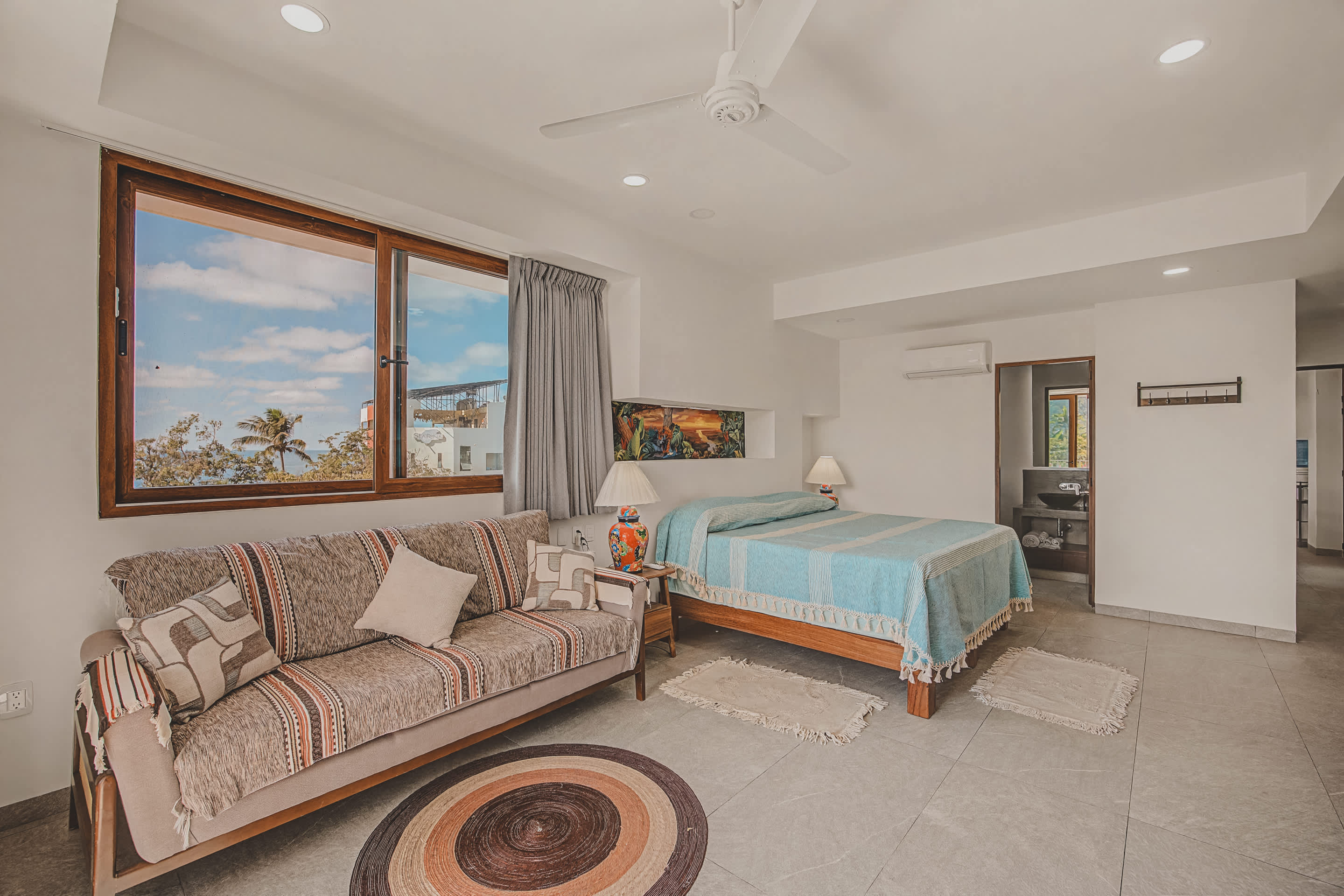 A bright bedroom features a patterned sofa and a bed with a light blue cover, set on light-colored tile flooring. A large window and sliding door offer views of the balcony and ocean.