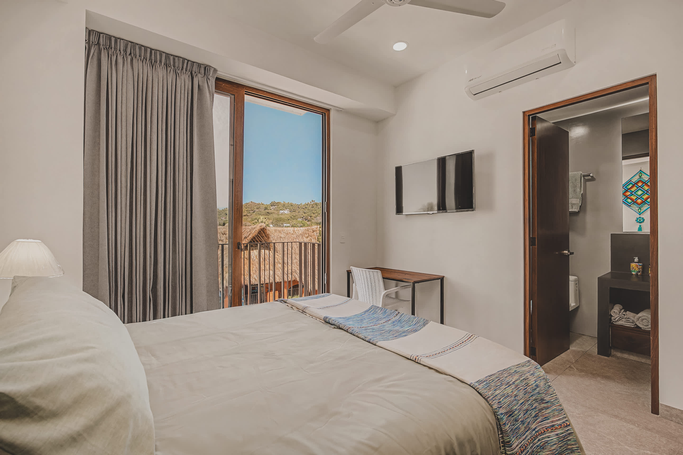 View of the bedroom from the foot of the bed, showcasing a wall-mounted TV, a small wooden desk with a white chair, and the open door to the ensuite bathroom. The balcony door with gray curtains offers a glimpse of the sunny outdoors.