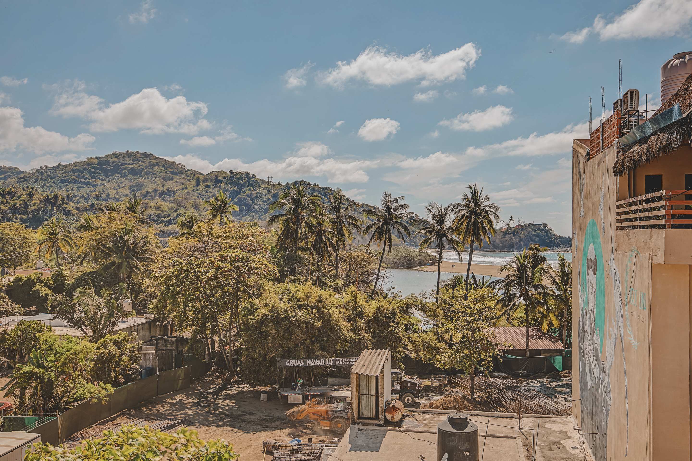 A panoramic view showcases a coastal landscape with a river meeting the ocean, surrounded by verdant hills and numerous palm trees. In the foreground, a dirt lot with a small shed and vehicles is visible.