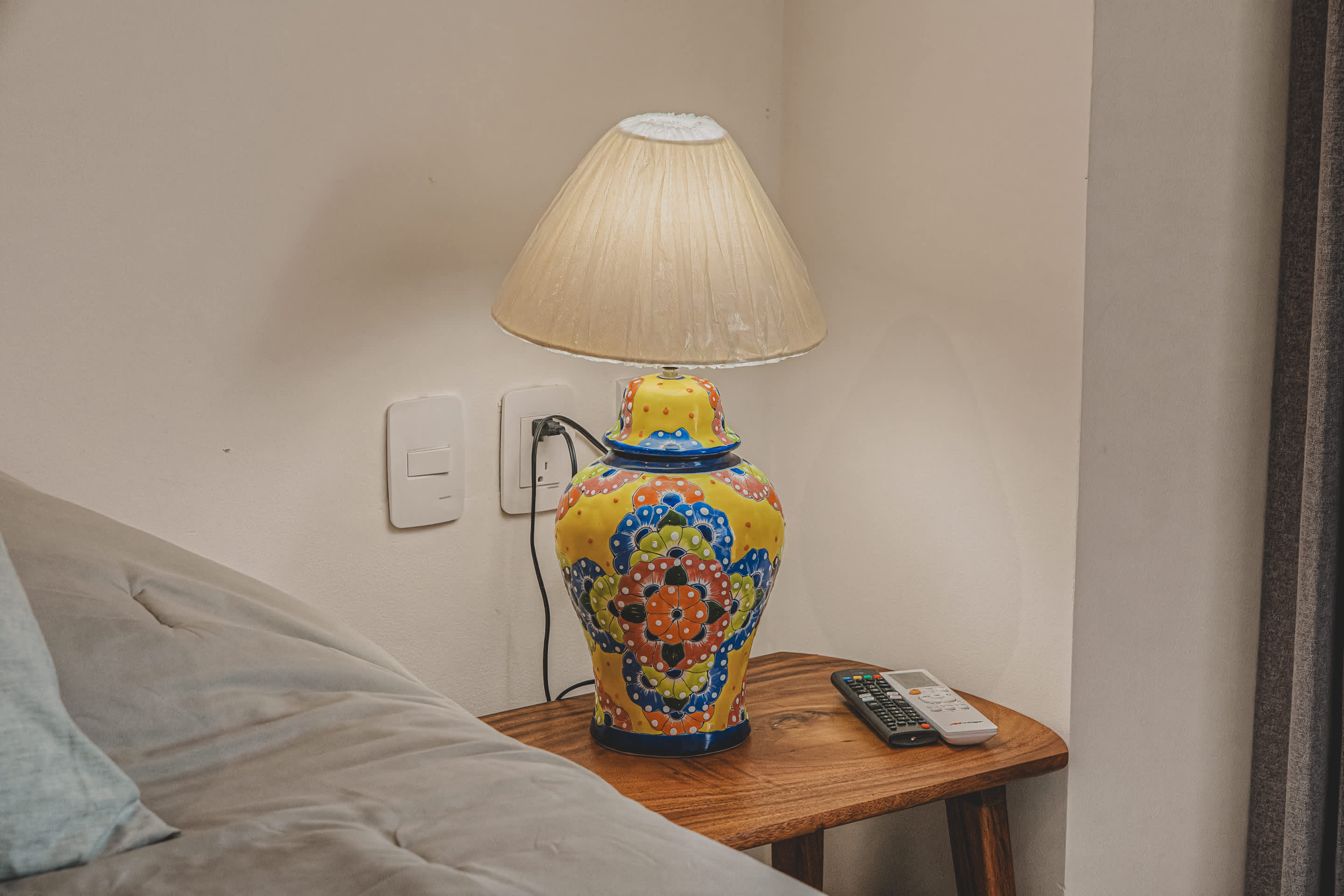 A vibrant, hand-painted ceramic table lamp with a yellow base and colorful floral patterns, sitting on a rustic wooden nightstand. Two remote controls are neatly placed beside the lamp.