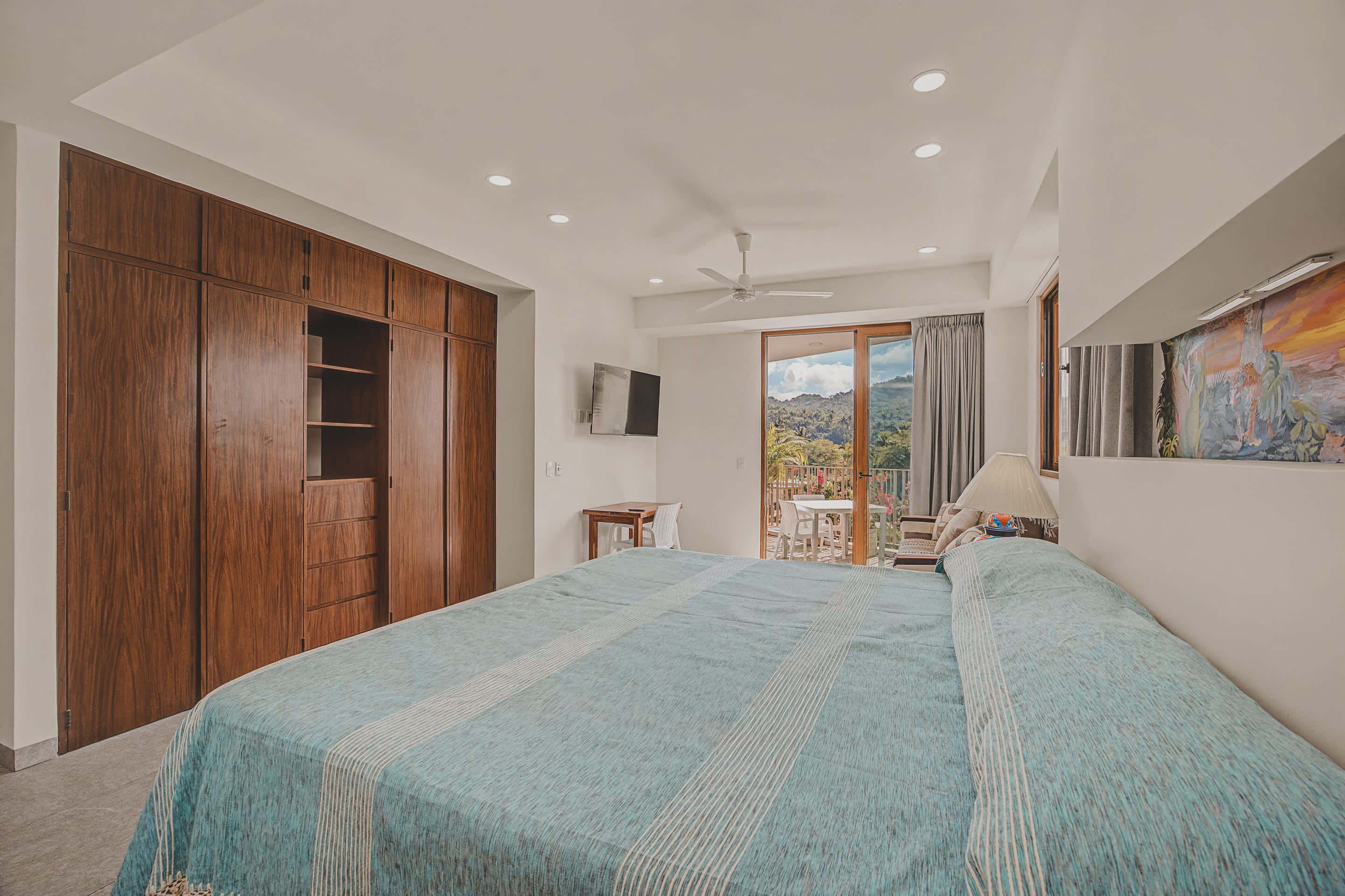 From the bed, a view reveals extensive built-in wooden wardrobes, a wall-mounted TV, and a sliding glass door providing access to the balcony and views of the surrounding landscape.