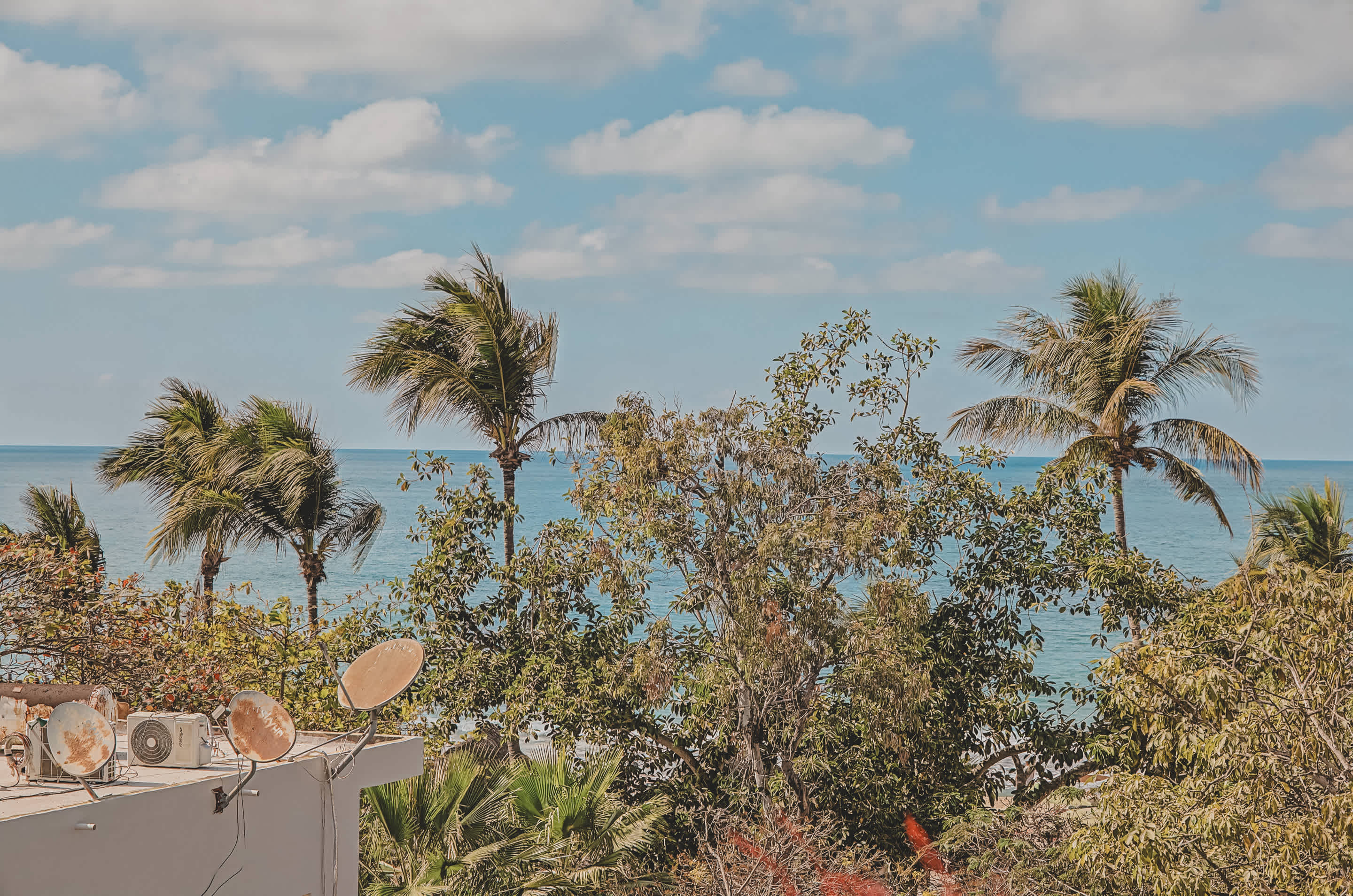 A vibrant ocean view from a balcony, featuring lush green palm trees against a clear blue sky with scattered white clouds. In the foreground, a white building with several satellite dishes is visible.