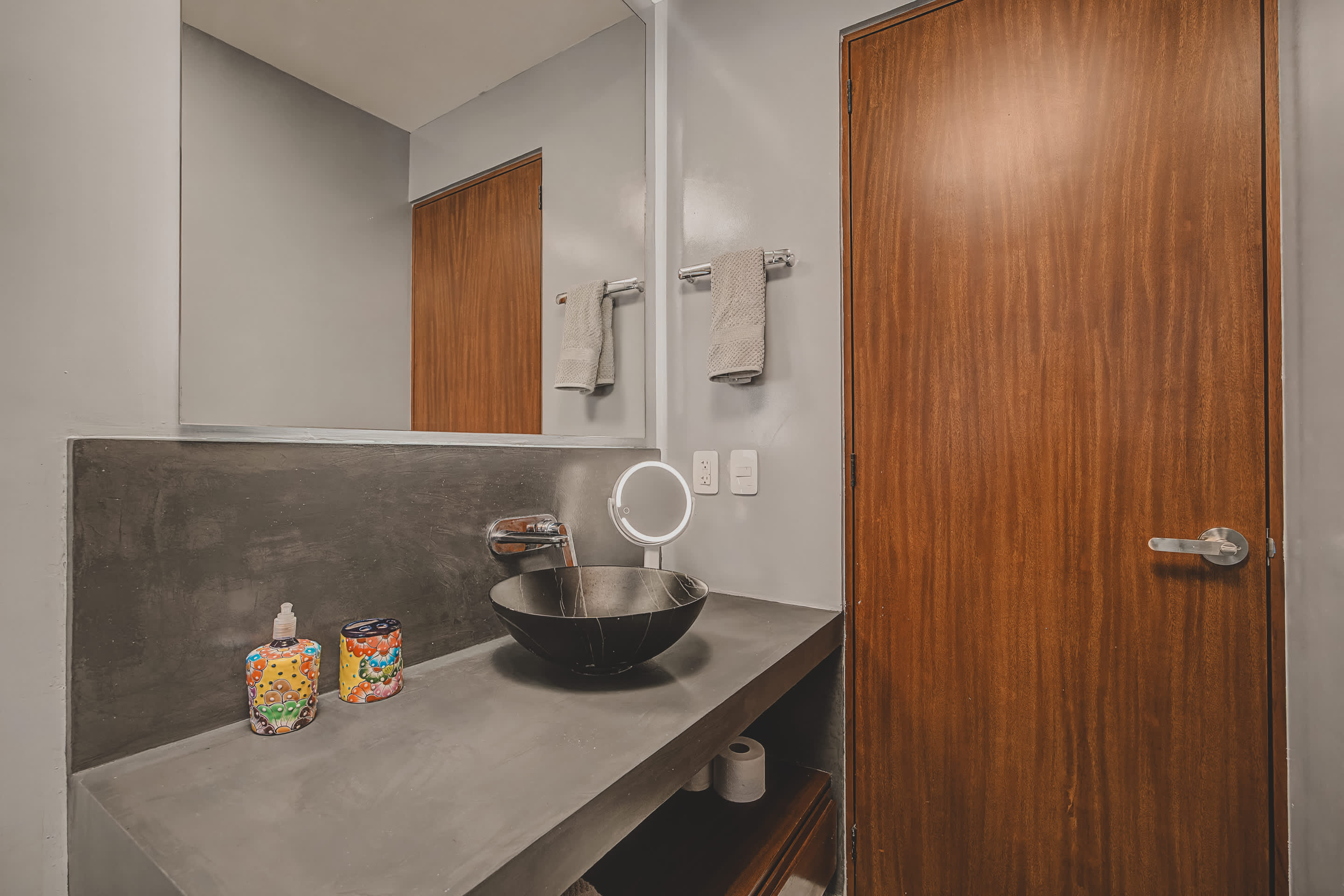 Contemporary bathroom featuring a dark concrete vanity with a black vessel sink and a large wall-mounted mirror. Colorful soap dispensers add a vibrant accent, and a warm wooden door is visible on the right.