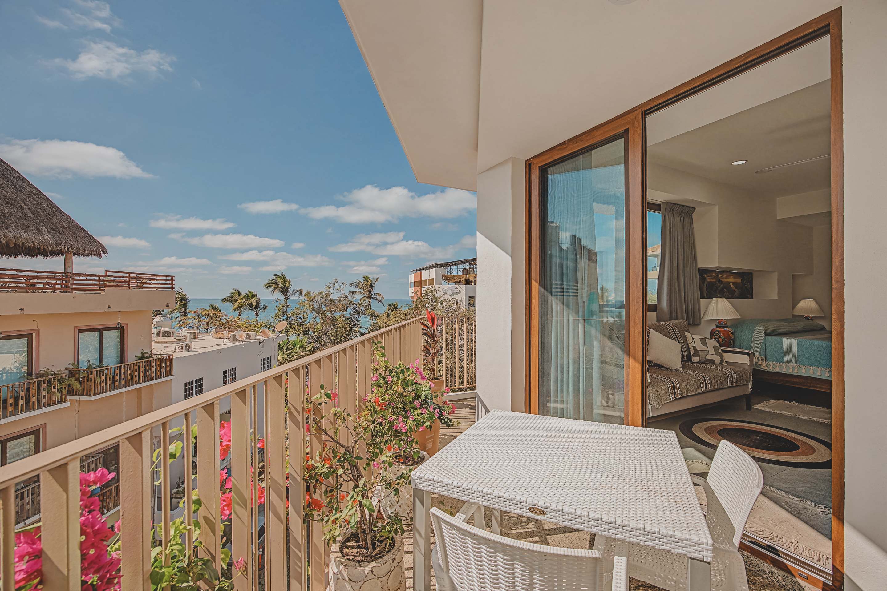 This balcony features a white outdoor dining set and potted plants, with a large sliding glass door providing direct access to an interior room. A clear ocean view is visible beyond the railing.