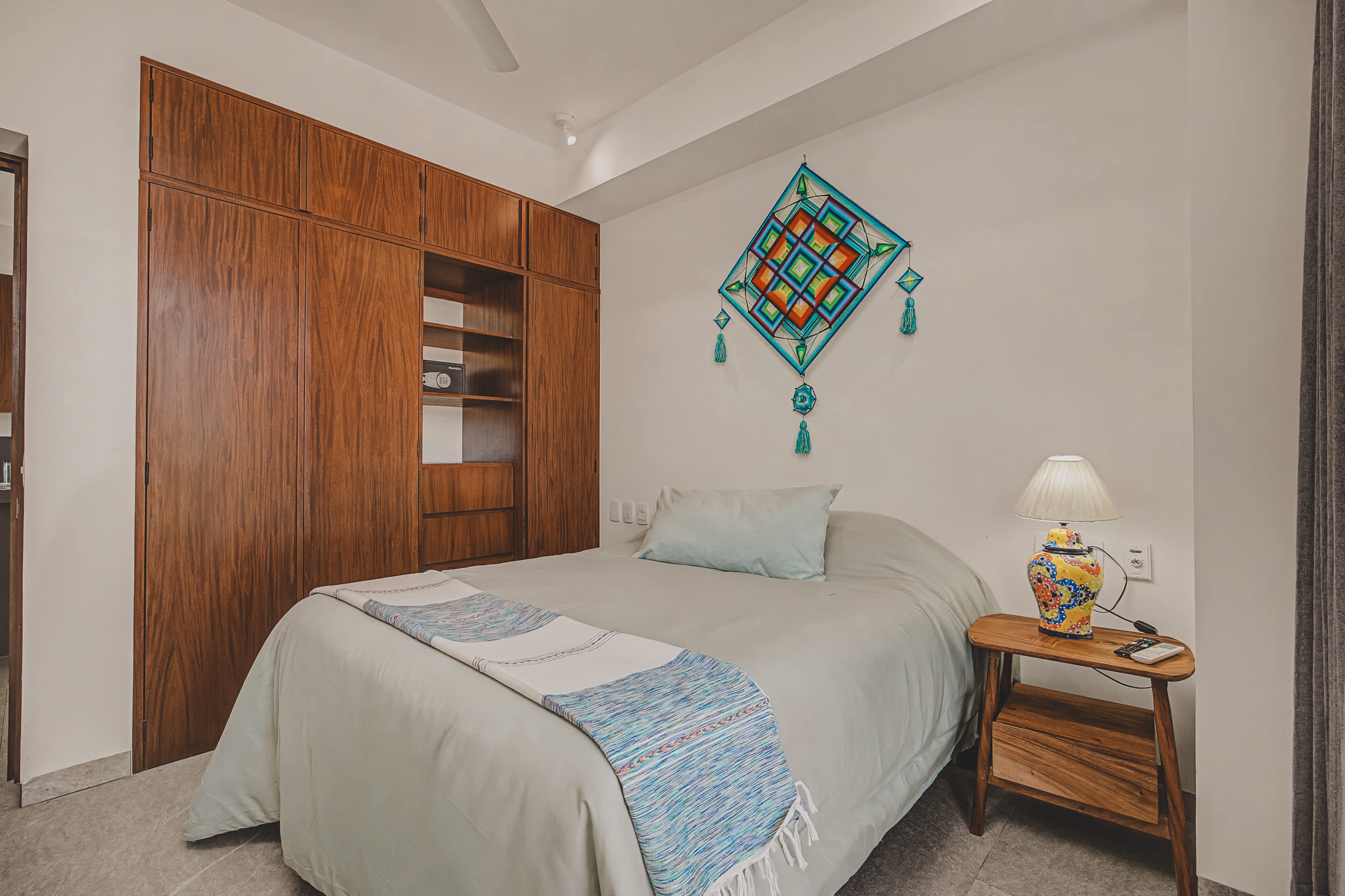 Cozy bedroom featuring a queen bed with light gray bedding and a blue and white patterned throw blanket. A wooden nightstand with a vibrant ceramic lamp sits beside the bed, complementing the colorful geometric wall art above the headboard.