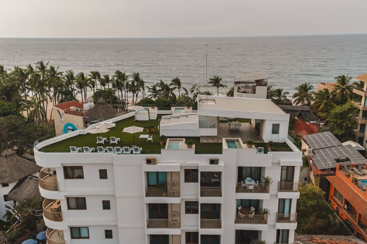 Aerial view of a modern white condominium building featuring a vibrant green rooftop terrace with lounge chairs, umbrellas, and small plunge pools. The property overlooks the ocean, with palm trees and other buildings visible along the coastline.
