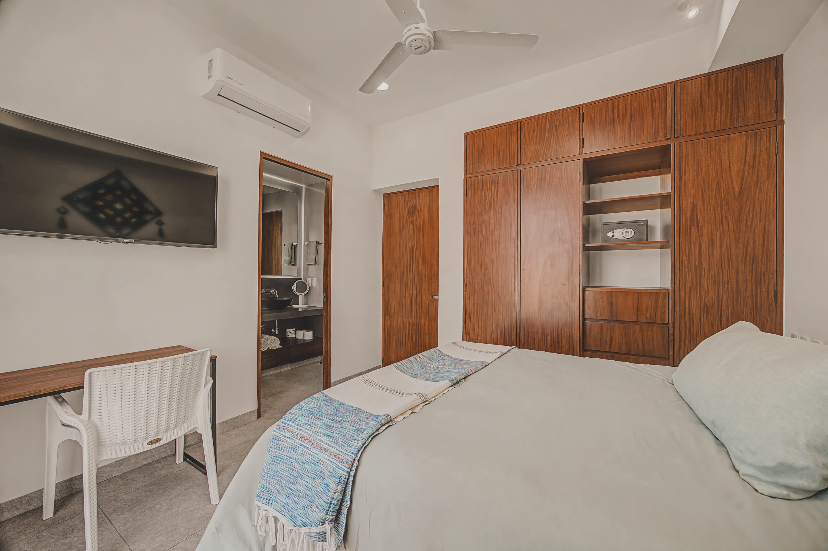 Bedroom featuring a queen bed, a large wooden built-in wardrobe with open shelving and a secure safe, and a wall-mounted TV. An open doorway leads directly into a modern bathroom with a dark vessel sink.