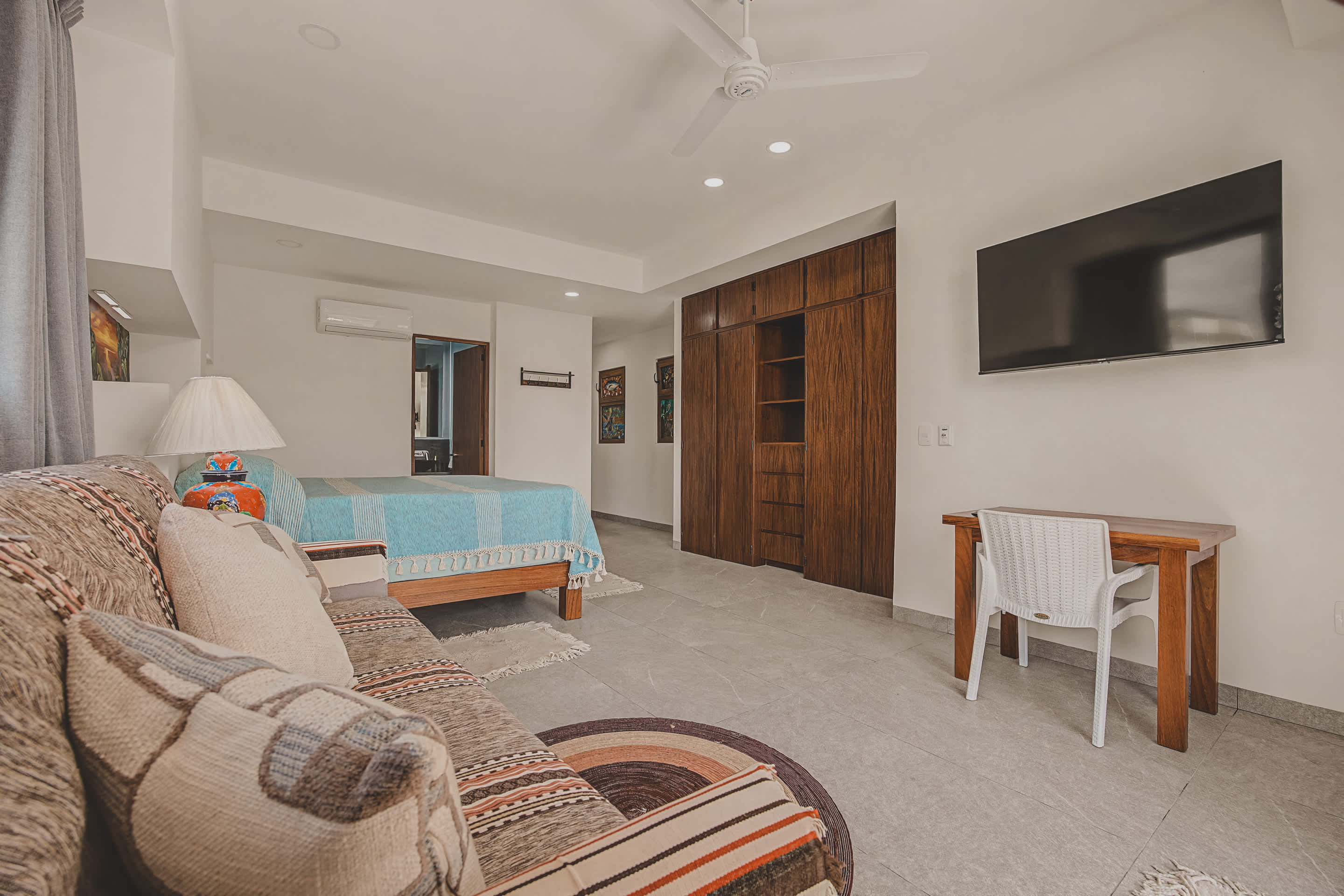 This bedroom includes a wall-mounted TV, a simple wooden desk with a white chair, and extensive built-in wooden wardrobes providing ample storage. Light-colored tile flooring continues throughout the space.