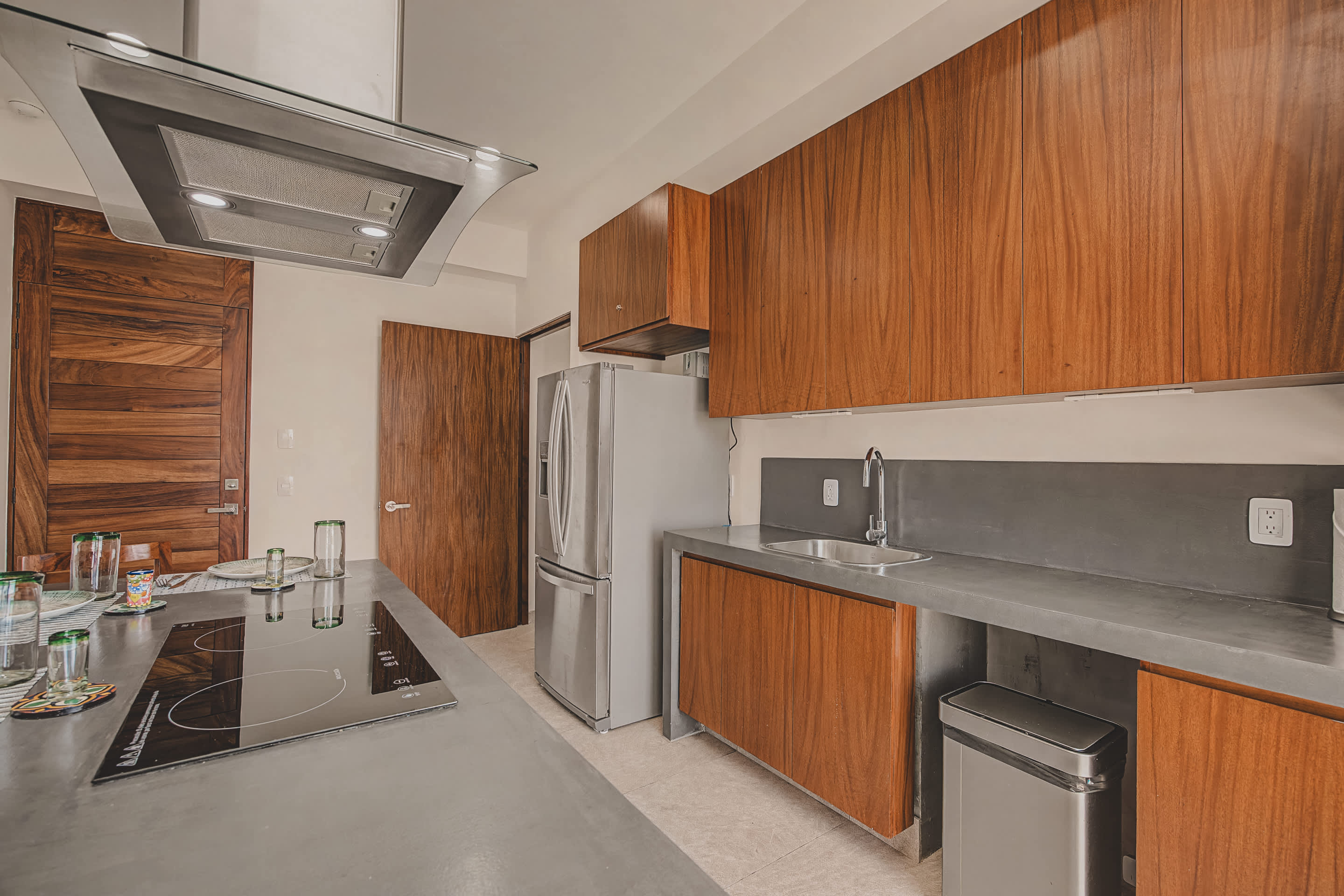 A close-up of the kitchen reveals a stainless steel refrigerator next to dark grey countertops with a sink, complemented by warm wooden cabinets and a sleek, modern design.