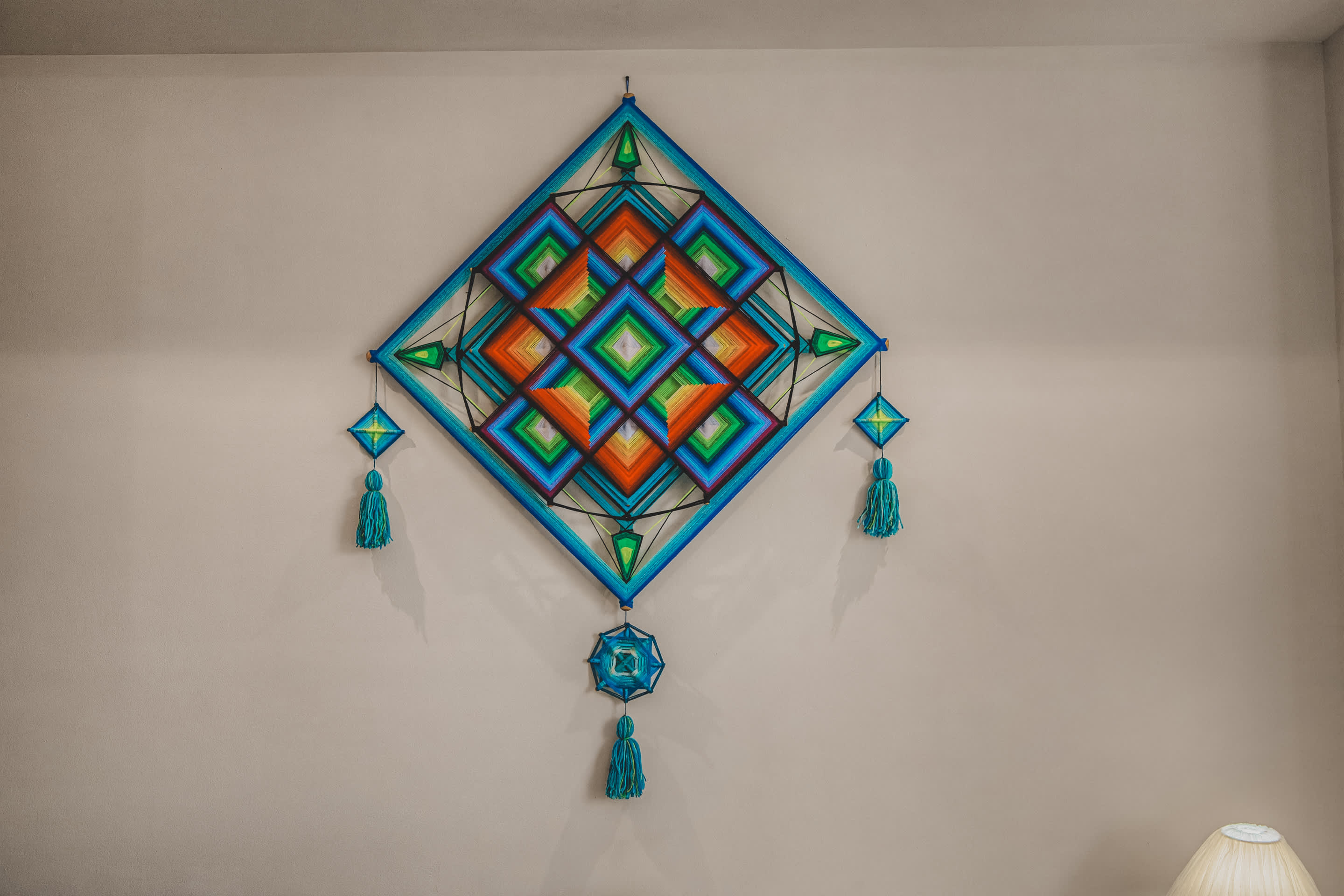 A striking, colorful yarn art piece, resembling a large diamond shape with intricate geometric patterns in shades of blue, green, orange, and red. It features decorative tassels hanging from its corners and bottom.
