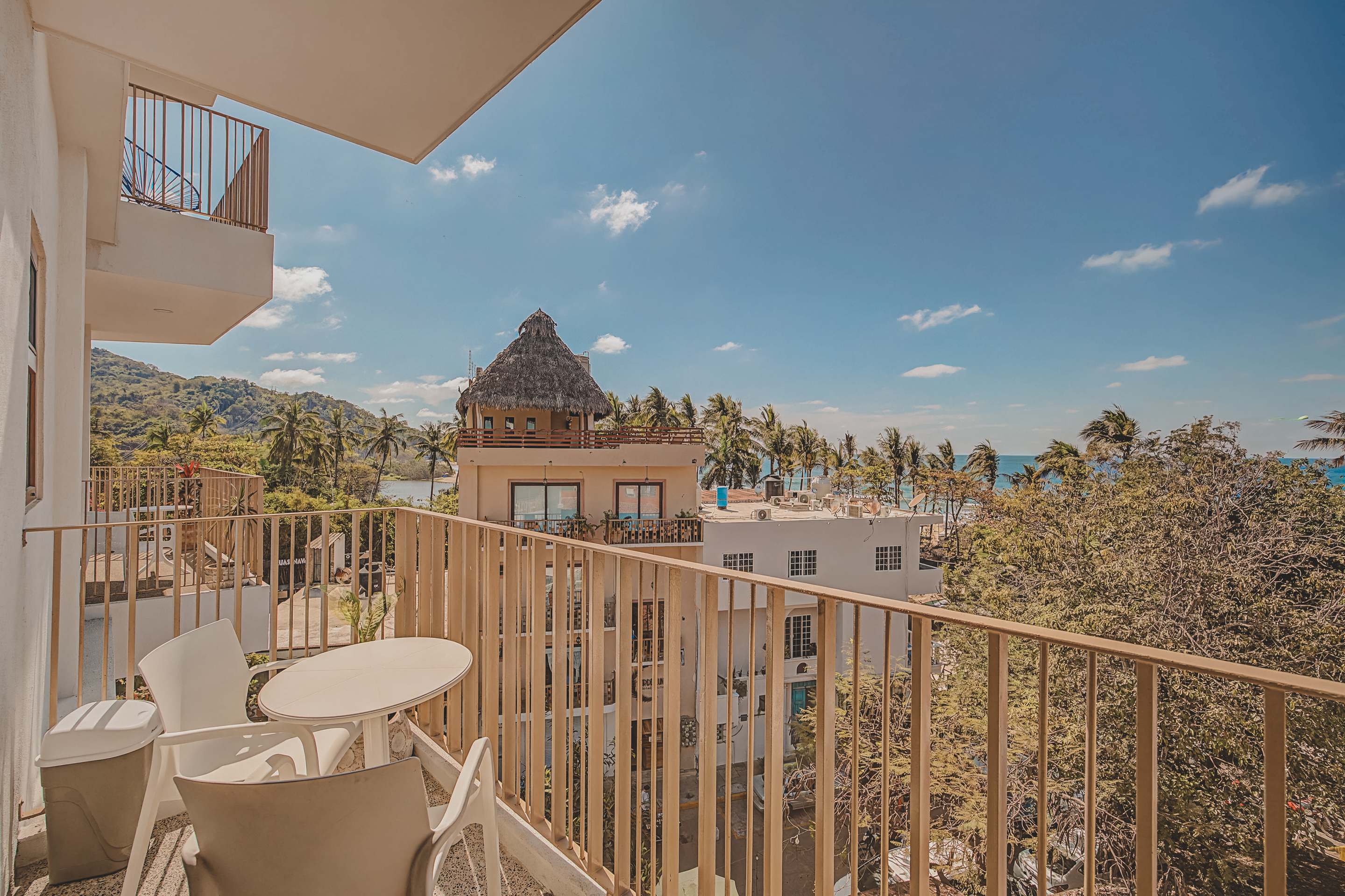 Balcony furnished with a small white table and chairs, offering a panoramic view of a charming coastal town with lush palm trees, various buildings, and the distant ocean. A distinctive thatched-roof building is prominent in the foreground.