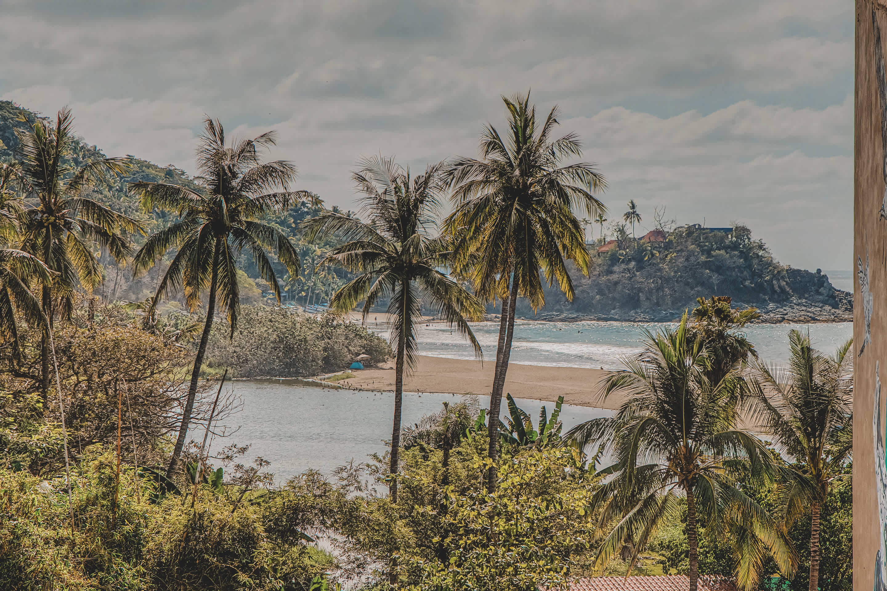 A scenic view of a tranquil bay with a sandy beach, surrounded by dense green foliage and numerous palm trees. Distant hills and a cloudy sky complete the picturesque coastal landscape.