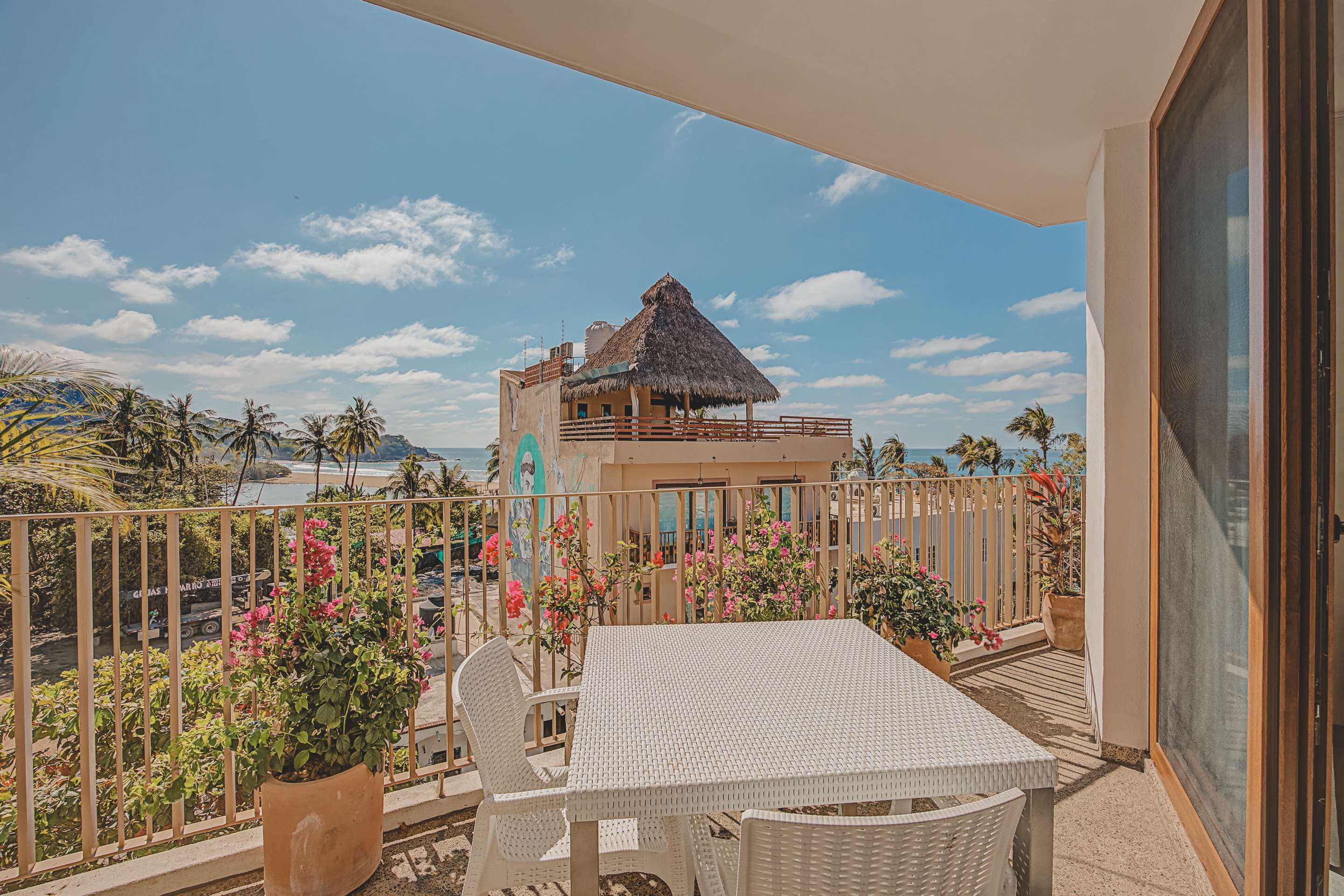 A bright balcony is furnished with a white outdoor dining table and chairs, adorned with blooming bougainvillea in terracotta pots. The view overlooks the ocean and nearby buildings, some with distinctive thatched roofs.