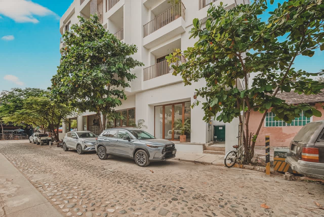 Street-level view of the multi-story white condominium building with multiple balconies, situated along a cobblestone street. Cars are parked curbside, and mature trees provide shade, contributing to a charming urban streetscape.