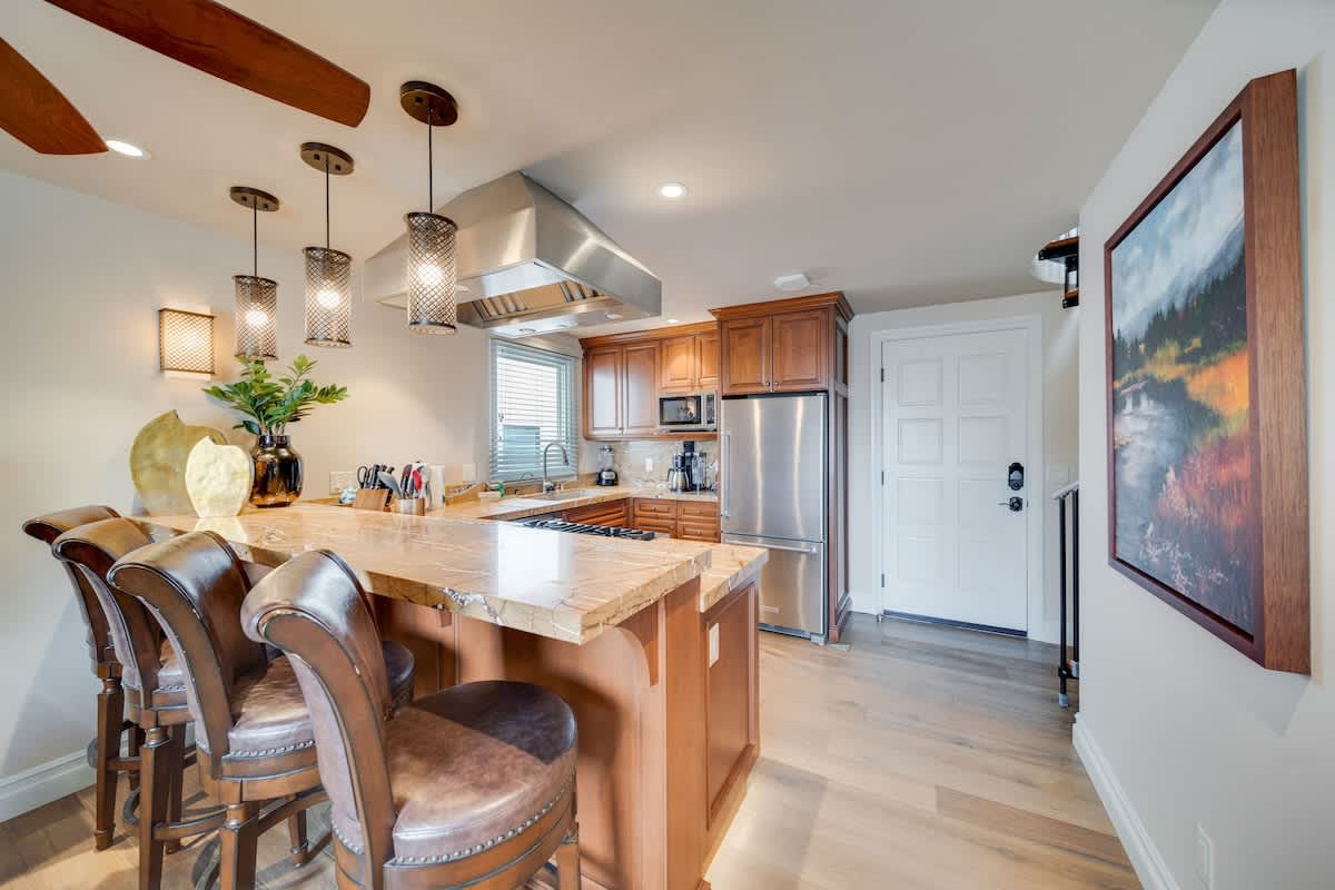View of the open-concept kitchen and living area, showcasing a large granite island with three leather bar stools, wood cabinetry, and stainless steel appliances. Pendant lights hang above the island, illuminating the space.