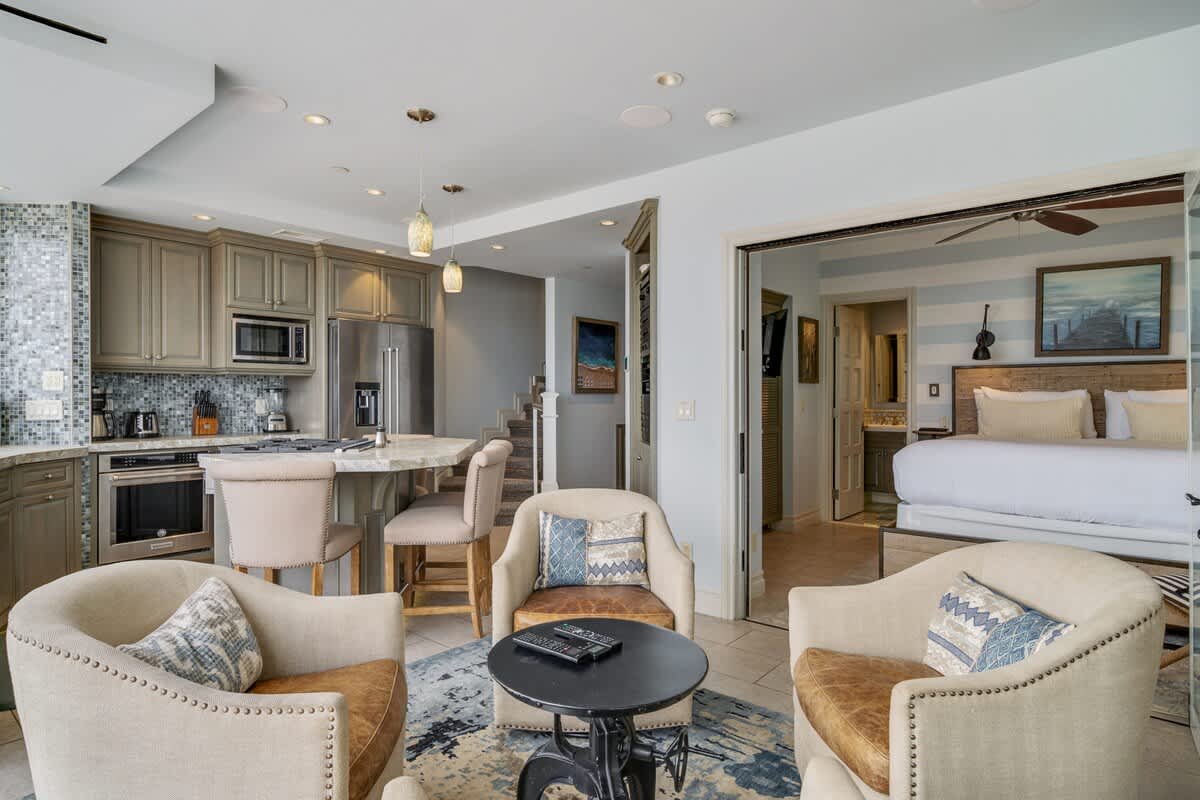 Open-concept living area with three upholstered armchairs and a small round table, connected to a modern kitchen, and an adjacent bedroom with a king-sized bed visible through an open doorway.