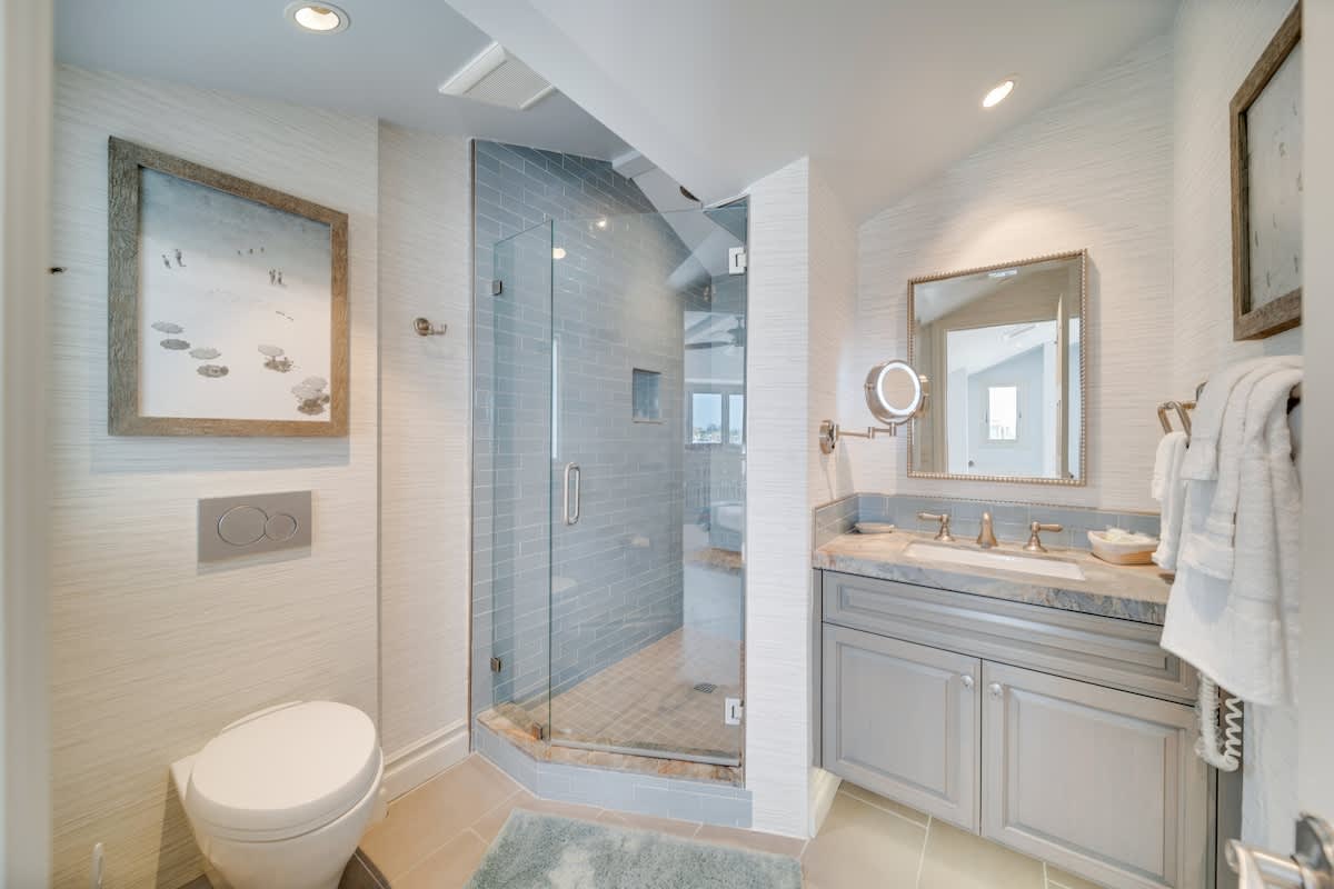 Modern bathroom featuring a glass-enclosed walk-in shower with blue subway tiles, a floating toilet, and a gray vanity with a stone countertop and undermount sink. The walls are covered in textured light wallpaper.