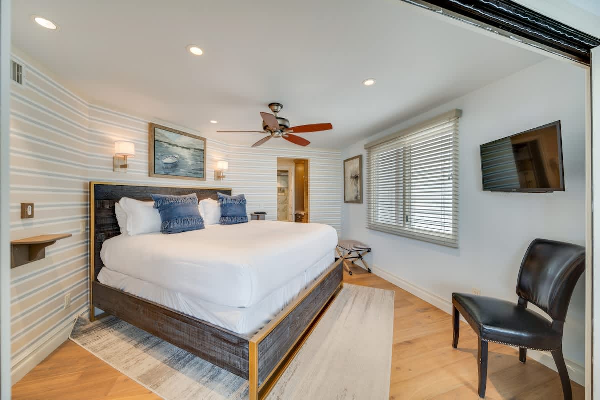 A spacious bedroom featuring a large bed with a dark wood and gold-accented frame, white bedding, and blue throw pillows, set against a striped accent wall. The room has light wood floors, recessed lighting, a ceiling fan, and a window with blinds.
