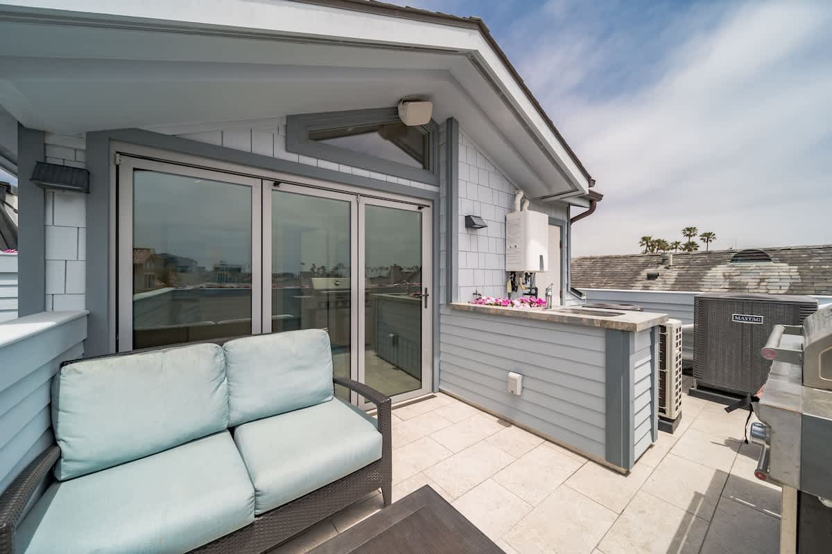 Spacious rooftop balcony with a comfortable outdoor sofa, a coffee table, and a built-in outdoor kitchen area with a BBQ grill. The light gray siding and white trim of the house are visible under a bright sky.
