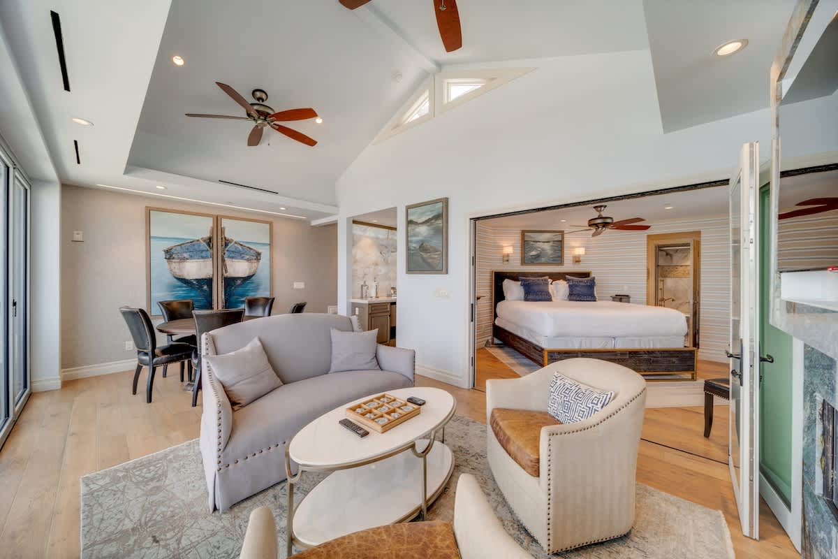 Bright living room with light hardwood floors, a gray sofa, and armchairs, leading to a dining area and an adjacent bedroom with a large bed, all featuring bay views through large glass doors.