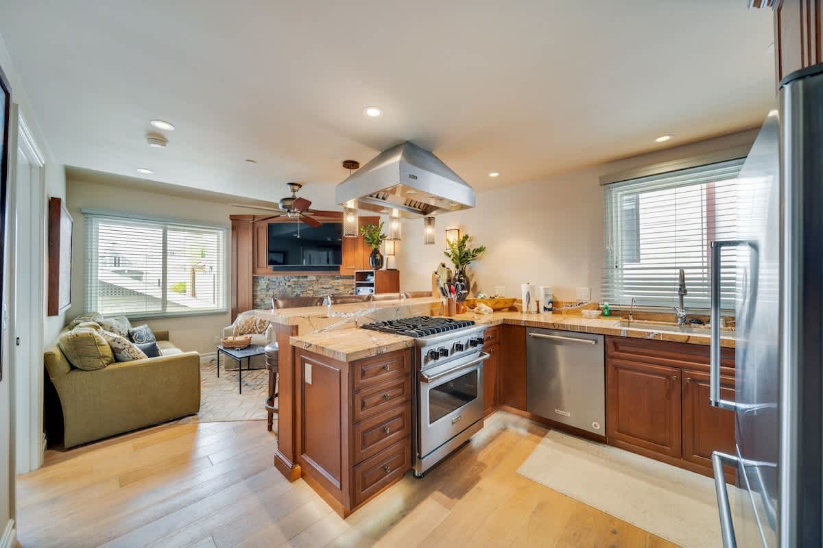 Open-concept kitchen with warm wood cabinetry, stainless steel appliances including a gas range and refrigerator, and a large island with a light-colored granite countertop. It flows into a living area with a sofa.