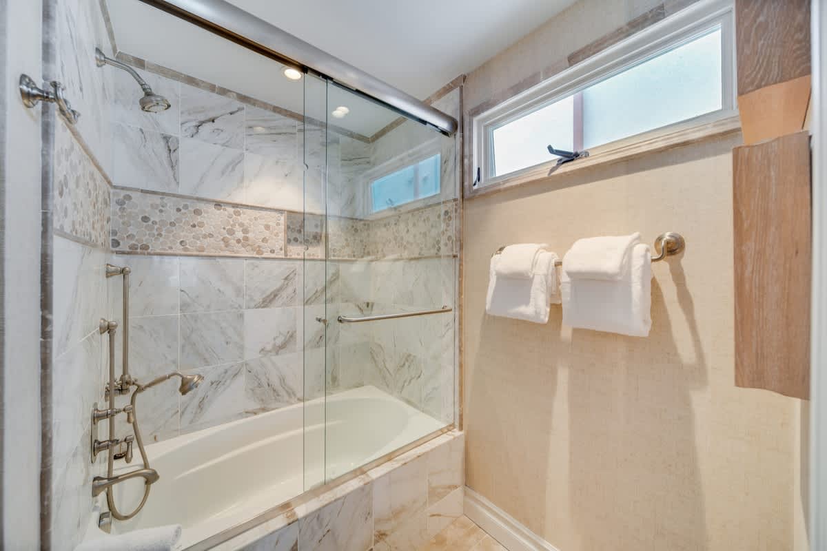 A clean shower and bathtub area features marble-patterned tiles, a glass sliding door, and a small window for natural light. Fresh white towels are neatly folded on a rack.