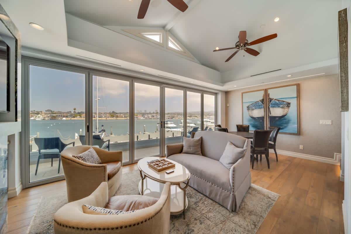 Elegant living room with light hardwood floors, a gray sofa, two armchairs, and a coffee table, adjacent to a dining area, all with expansive bay views through floor-to-ceiling sliding glass doors.