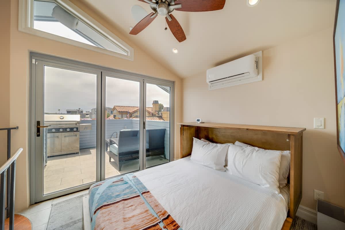 Cozy bedroom with a white bed, a wall-mounted air conditioning unit, and large sliding glass doors that open to a balcony with a BBQ grill. A wooden ceiling fan complements the vaulted ceiling.