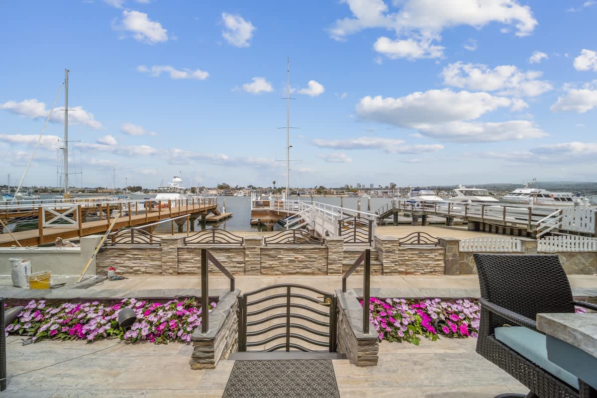 A stone patio area with a decorative metal gate, vibrant pink flowers, and outdoor seating, offering direct views of the bay with multiple boats and docks under a bright sky.