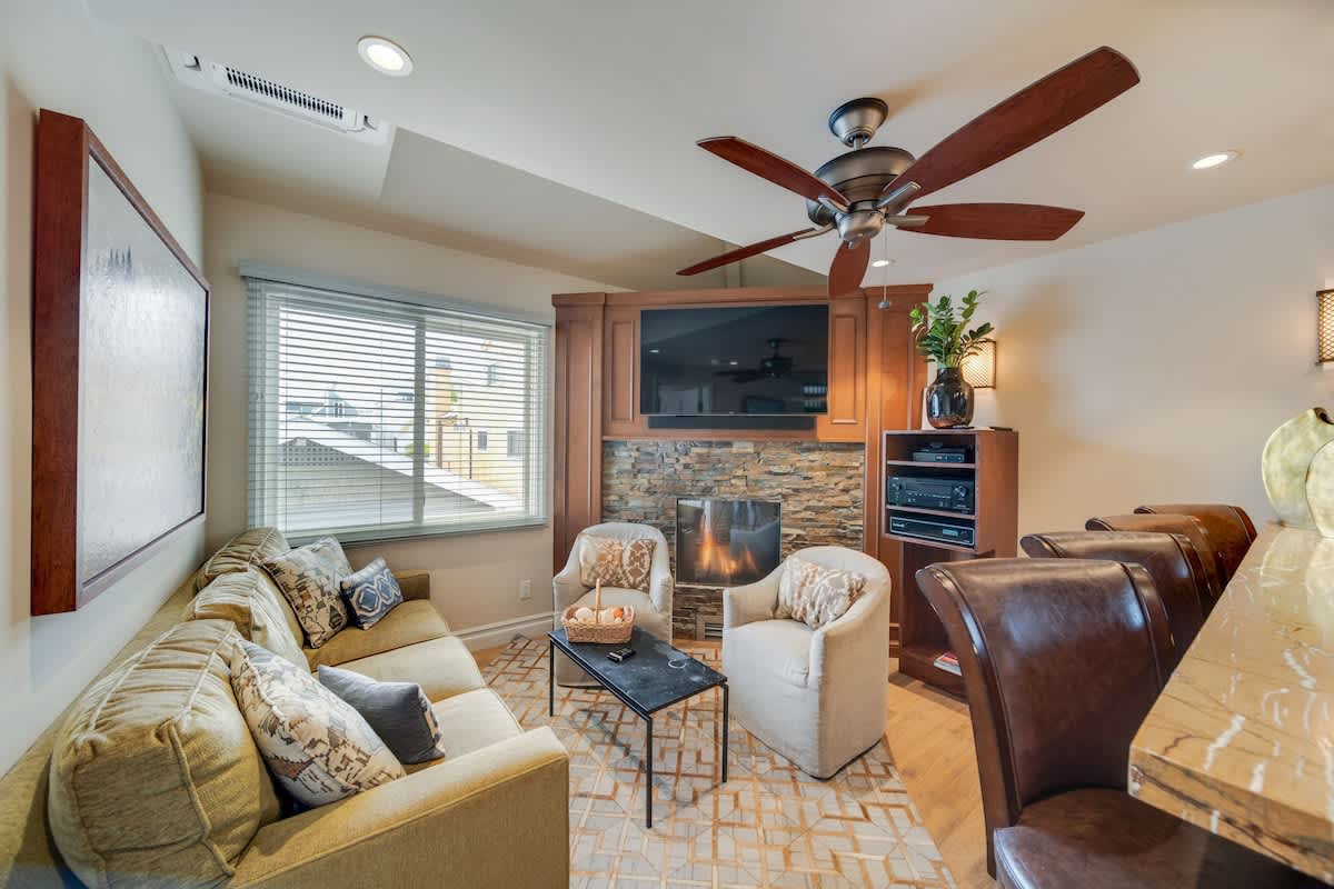 Cozy living room with a comfortable olive green sofa, two upholstered armchairs, and a stone-faced fireplace with a wall-mounted TV above it. A ceiling fan and large window with blinds complete the space.