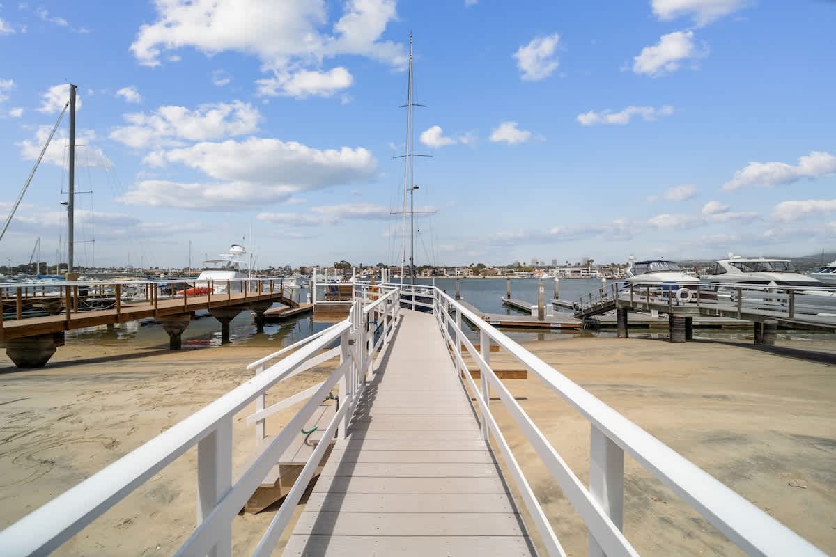 A long wooden walkway with white railings extends over a sandy beach towards multiple boat docks and yachts in a sunny bay. The water is calm under a blue sky with scattered clouds.